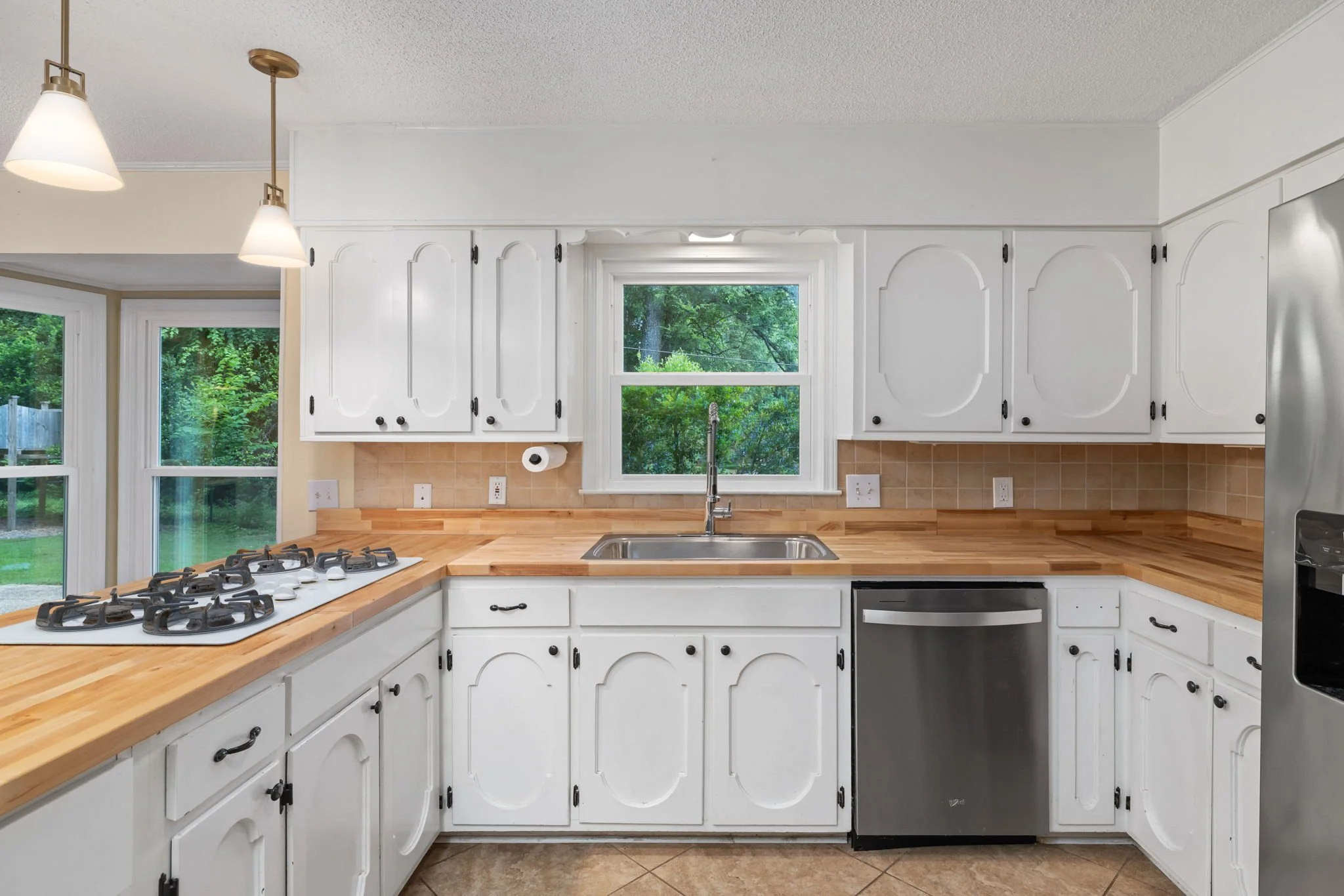 A bright kitchen with white cabinets and a wooden countertop, featuring a double window looking outside at greenery, a gas stove, a sink, and a dishwasher.