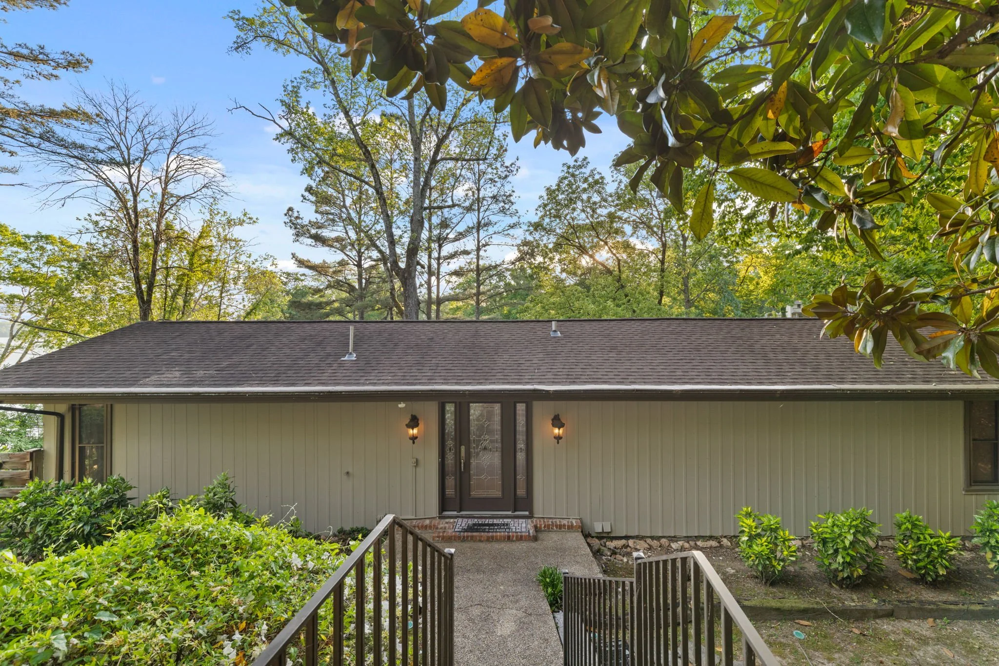 View of a house with a backyard, featuring a stairs leading down to a garden with bushes, and a roof with two vents. Tall trees with green and some yellow leaves are visible in the background under a blue sky.