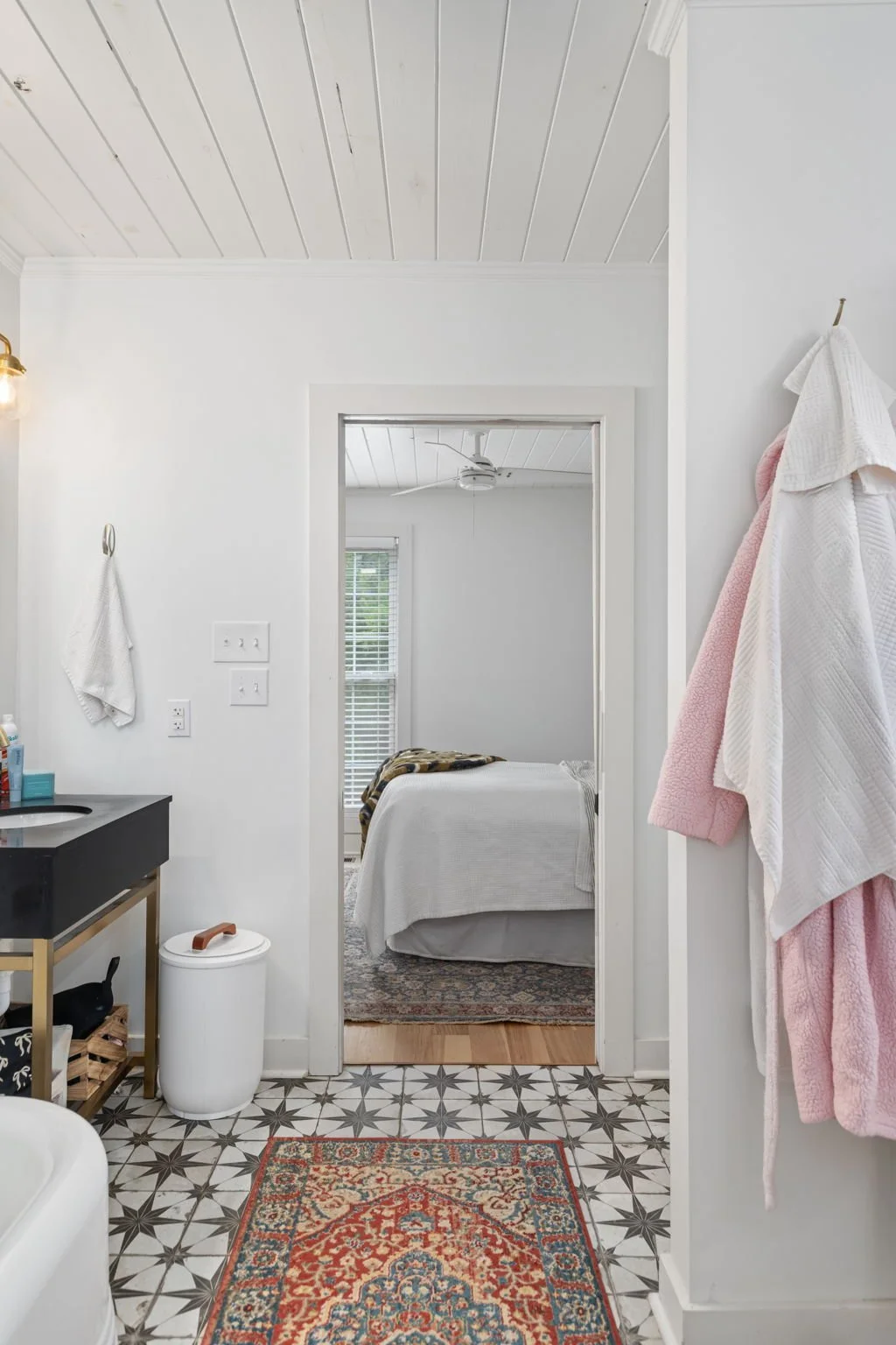 View of a bedroom seen through an open doorway, with a bed, window with blinds, ceiling fan, and a patterned rug, framed by a white wall with hanging towels and a patterned floor in the foreground.