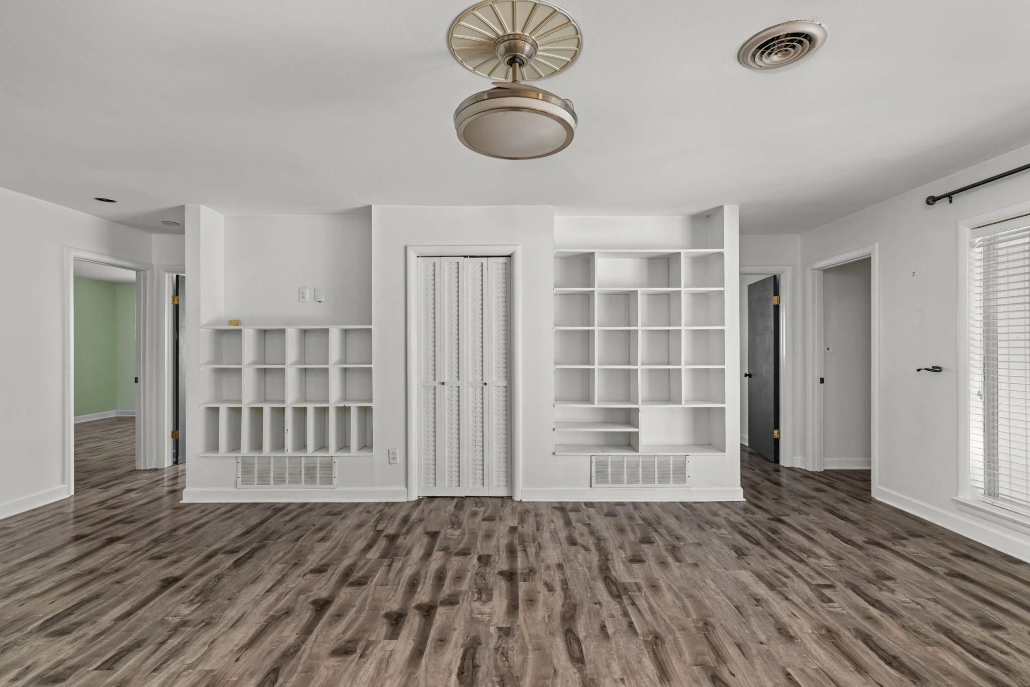 An empty room with white walls, wood-patterned flooring, a built-in white bookshelf, and a ceiling light fixture.