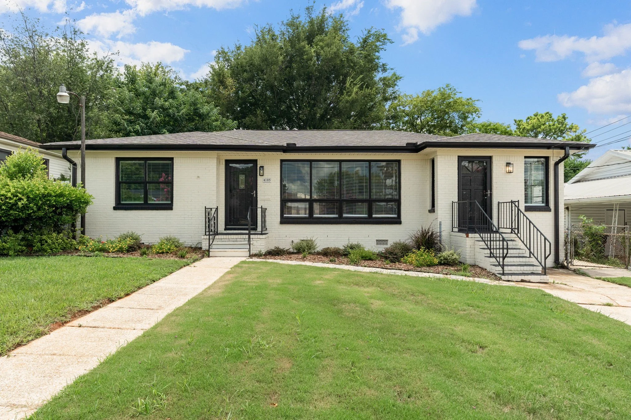 Front view of a white, single-story house with black window frames and stairs leading to two black doors, surrounded by a green lawn and garden with bushes, under a blue sky with a few clouds.