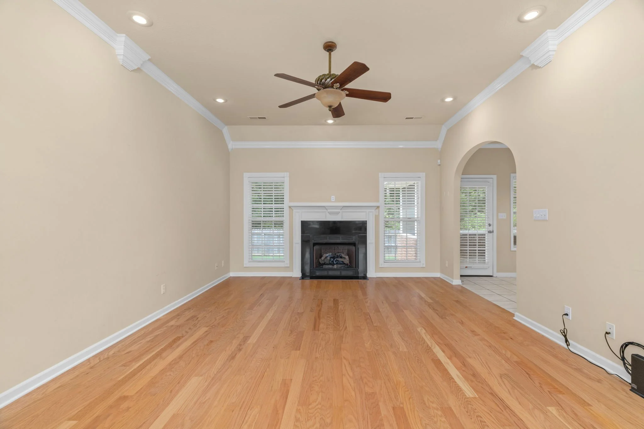 Empty living room with hardwood floors, beige walls, white crown molding, a ceiling fan, a fireplace, three windows with blinds, and an arched doorway leading to another room.