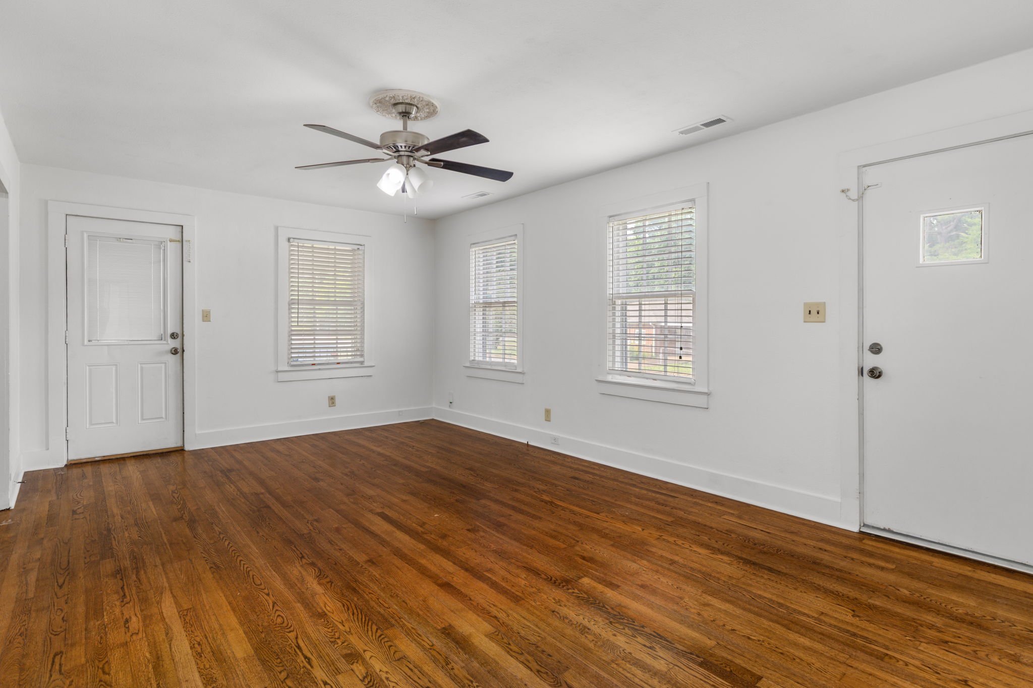 An empty room with hardwood flooring, white walls, several windows with blinds, a ceiling fan with lights, and a white door.