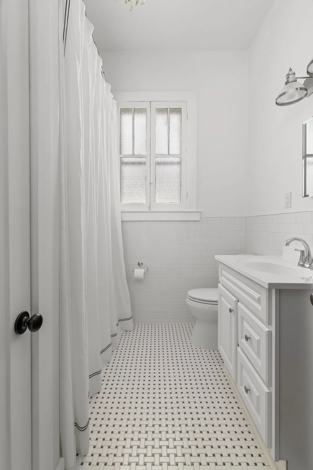 A small white bathroom with a window, a toilet, a white vanity with a sink, a mirror, a light fixture, white subway tile halfway up the wall, and a patterned tile floor.
