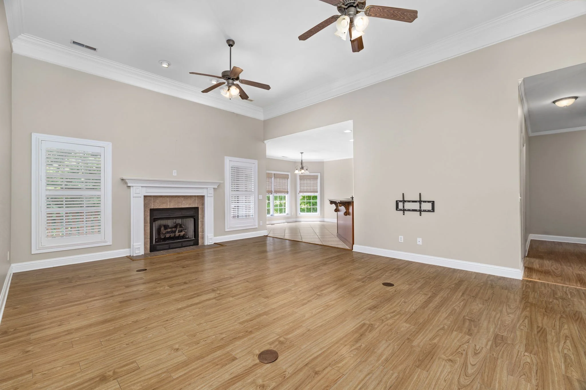 Empty living room with beige walls, white crown molding, wooden floors, a fireplace, multiple windows with blinds, ceiling fans, and a mounted TV bracket.