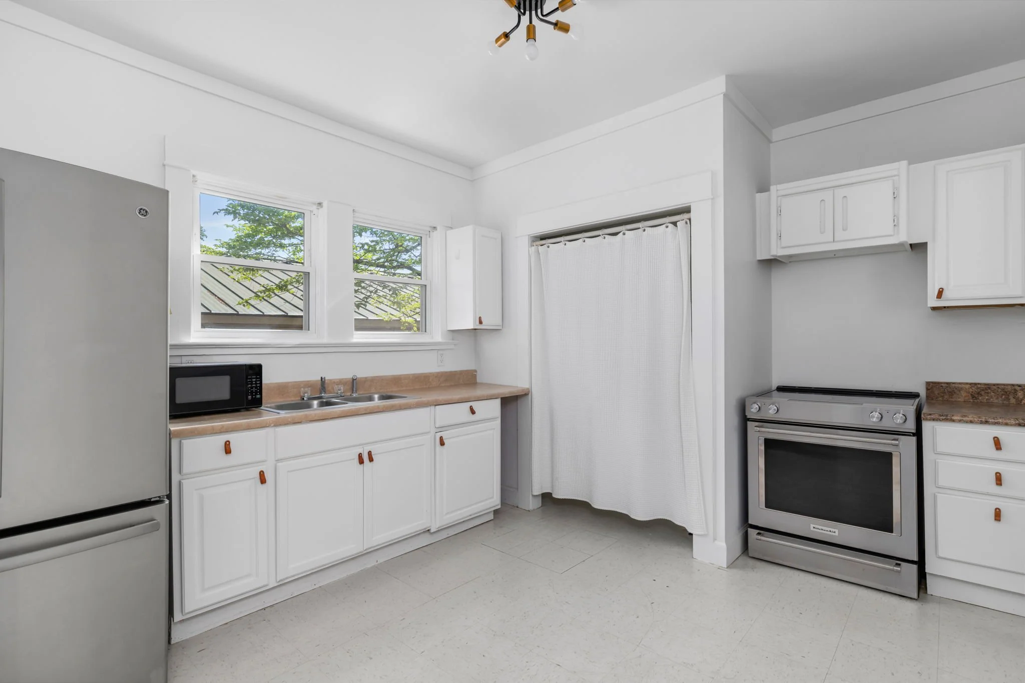 A kitchen with white cabinets, beige countertop, stainless steel refrigerator, microwave, and stove, with two windows showing trees outside and a ceiling light fixture.