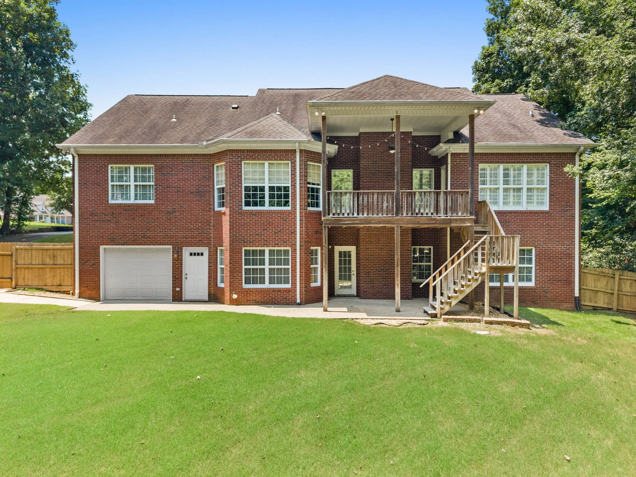 Back view of a brick house with multiple windows, a garage, and an elevated wooden deck with stairs, surrounded by a green lawn and tall trees.