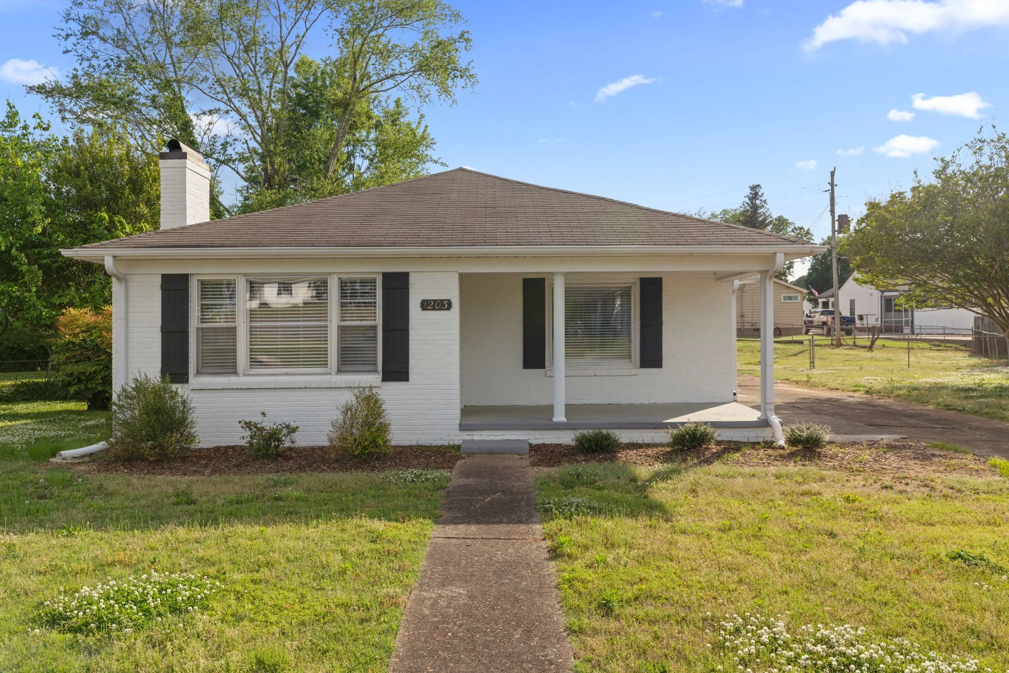 Front view of a white brick house with black shutters, a small front porch, and a brown shingled roof, surrounded by green grass, bushes, and trees under a blue sky with a few clouds.