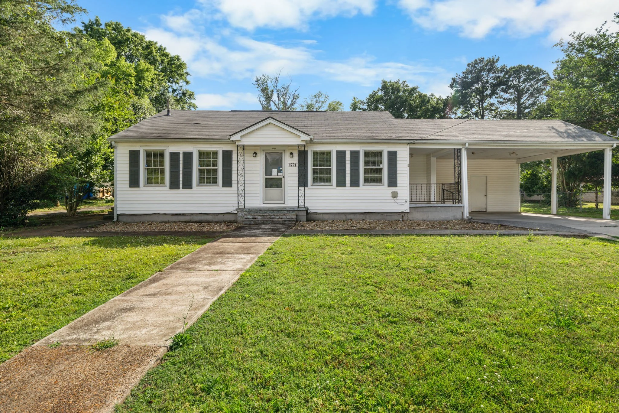 White house with a covered front porch, black shutters, and a front pathway leading to steps, surrounded by a green lawn and trees, under a partly cloudy sky.