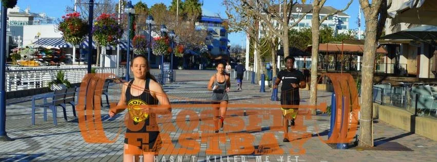 Three women and one man running outdoors on a paved walkway, with trees, benches, and buildings in the background, in a park or urban area.