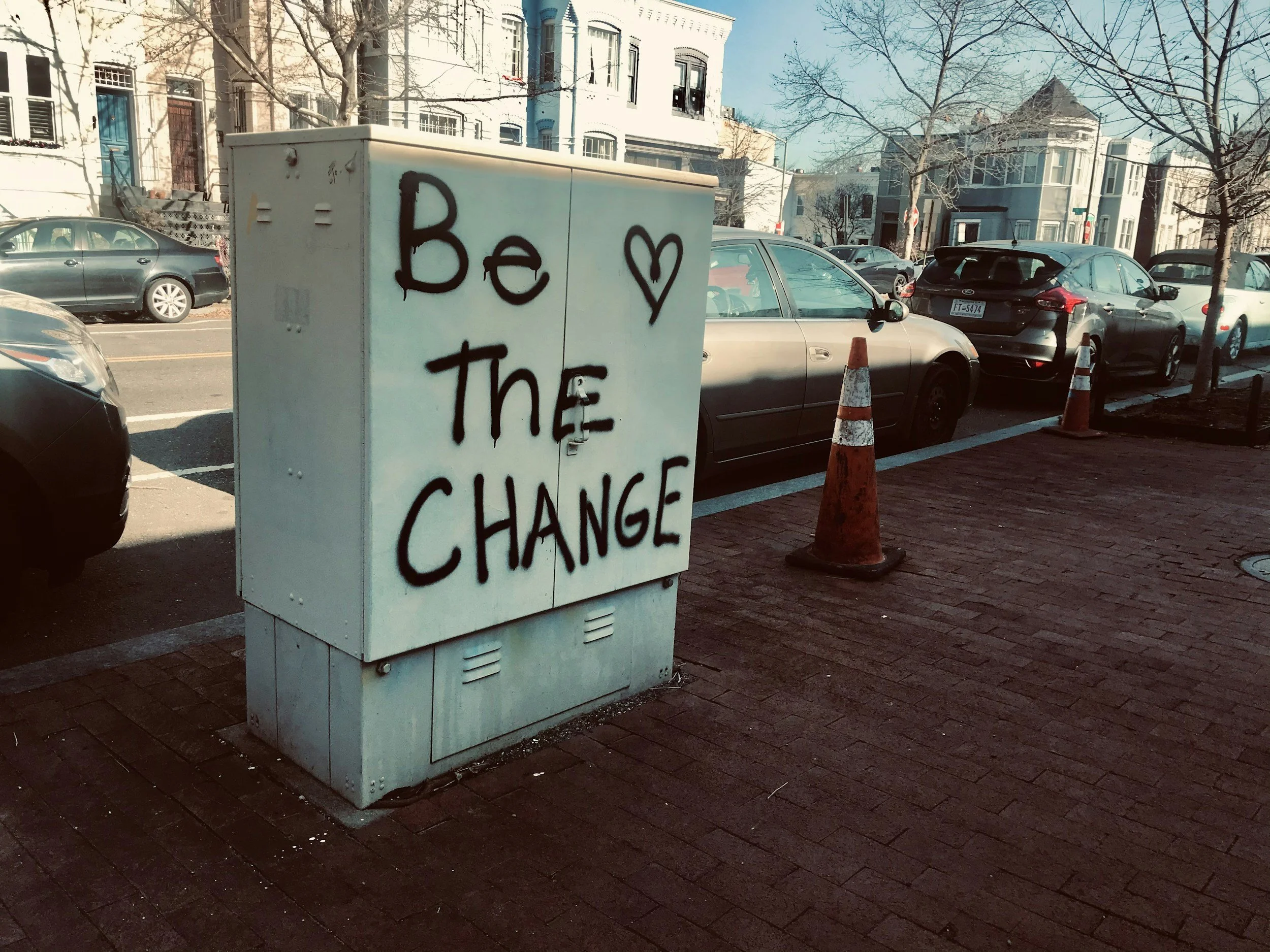 A white utility box on a brick sidewalk with black spray-painted words "Be The Change" and a heart symbol. Parked cars and residential buildings are visible in the background, with clear skies and leafless trees.
