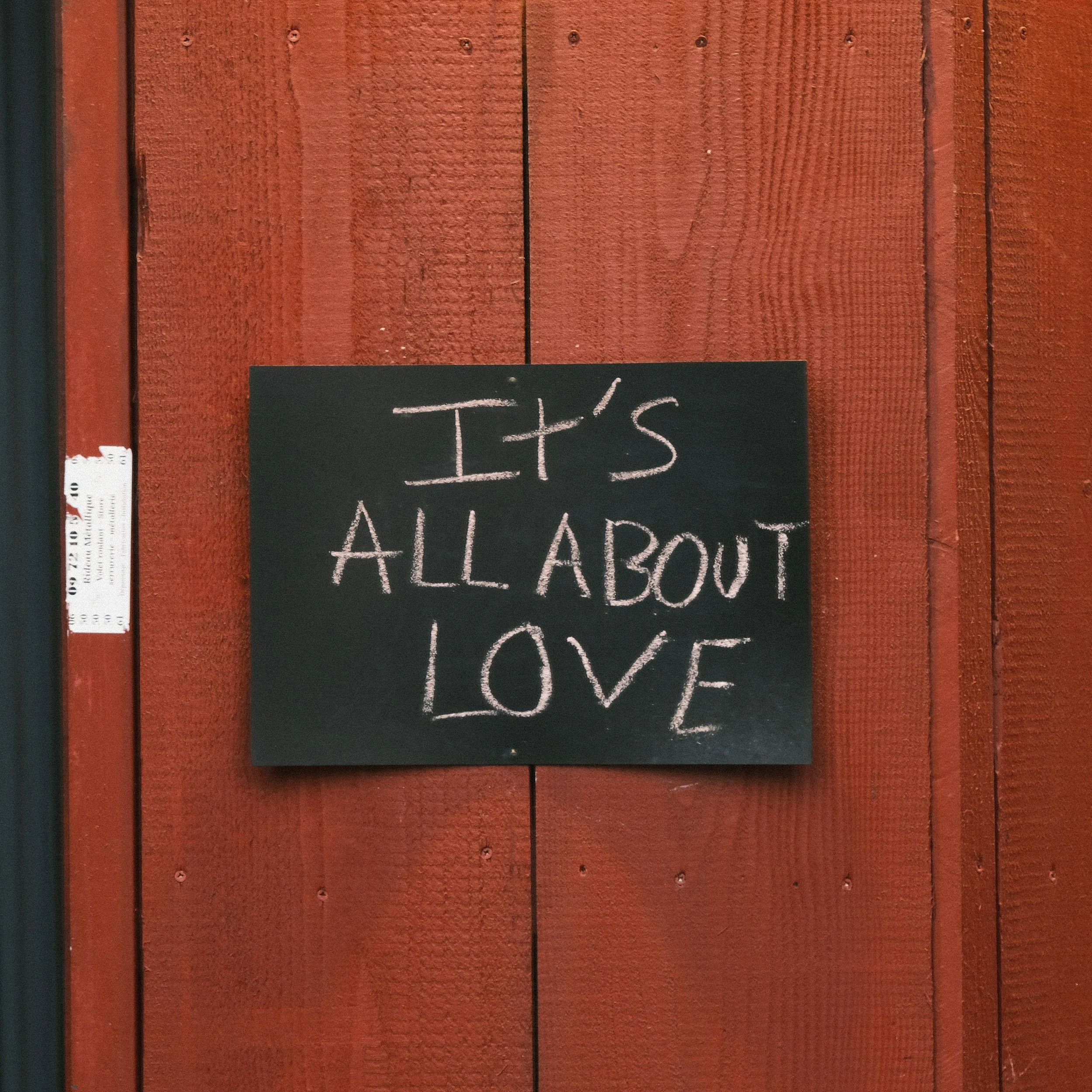 A black chalkboard sign with the message 'It's all about love' written in white chalk, hanging on a red wooden wall.