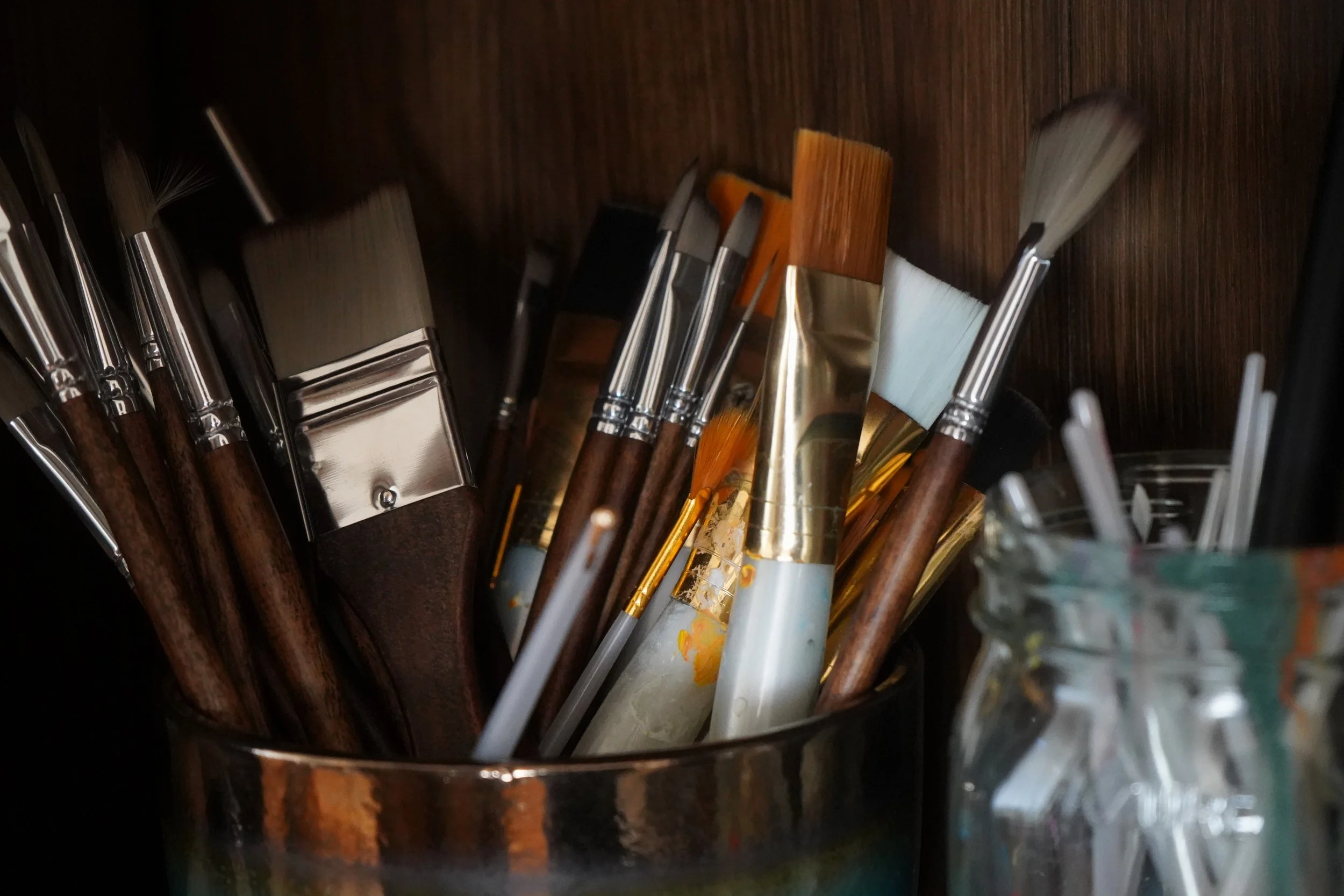 Photograph of an assortment of artist's paintbrushes in a studio container beside painting tools.