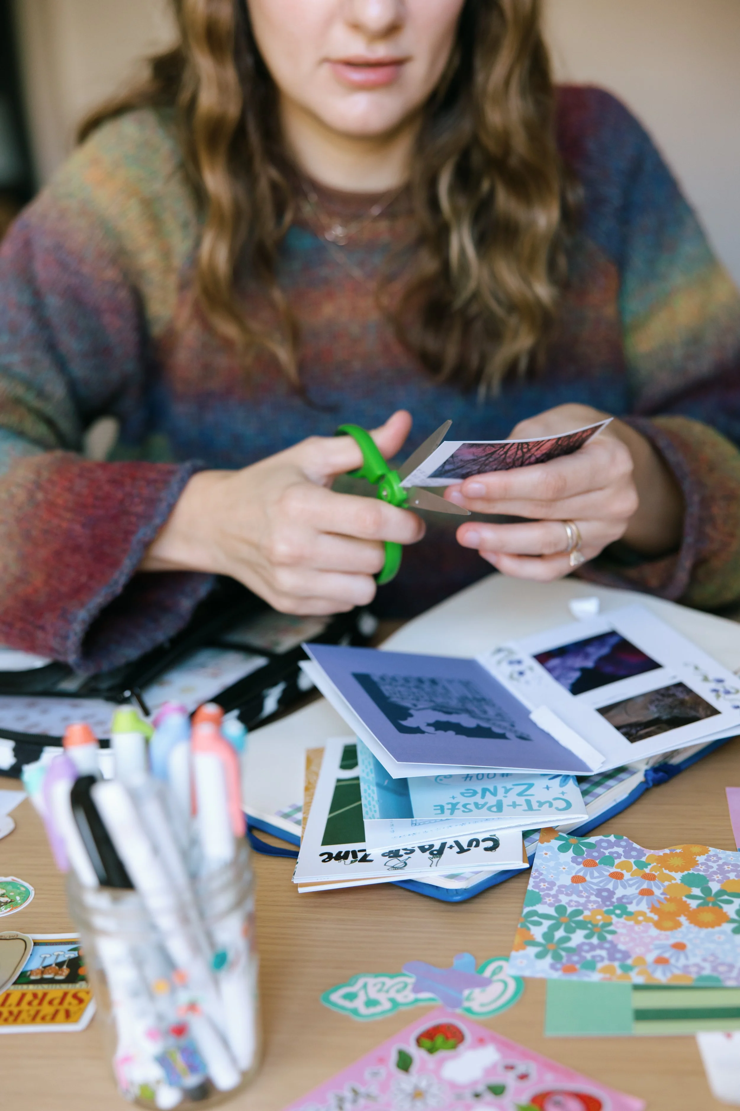 A person with wavy hair wearing a multicolored sweater is cutting a printed photo with a pair of scissors. The table is cluttered with colorful papers, stickers, and craft supplies, including markers in a jar and various printed materials.
