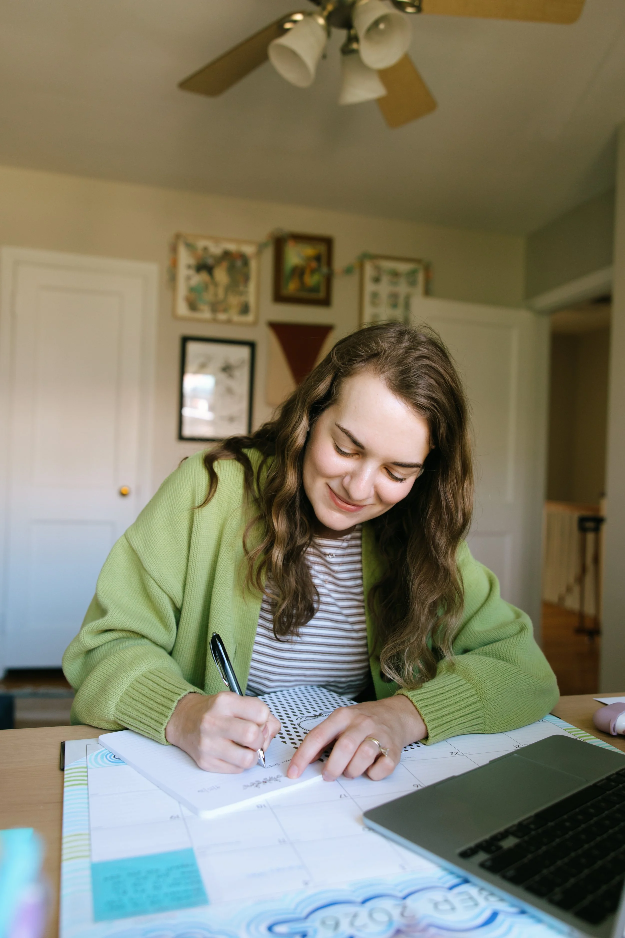 A young woman with long curly brown hair, wearing a green cardigan and striped shirt, is sitting at a desk, smiling, and writing in a notebook with a black pen. The desk has various papers, a laptop, and a pink tape dispenser. The background shows a beige wall with framed artwork and a ceiling fan.