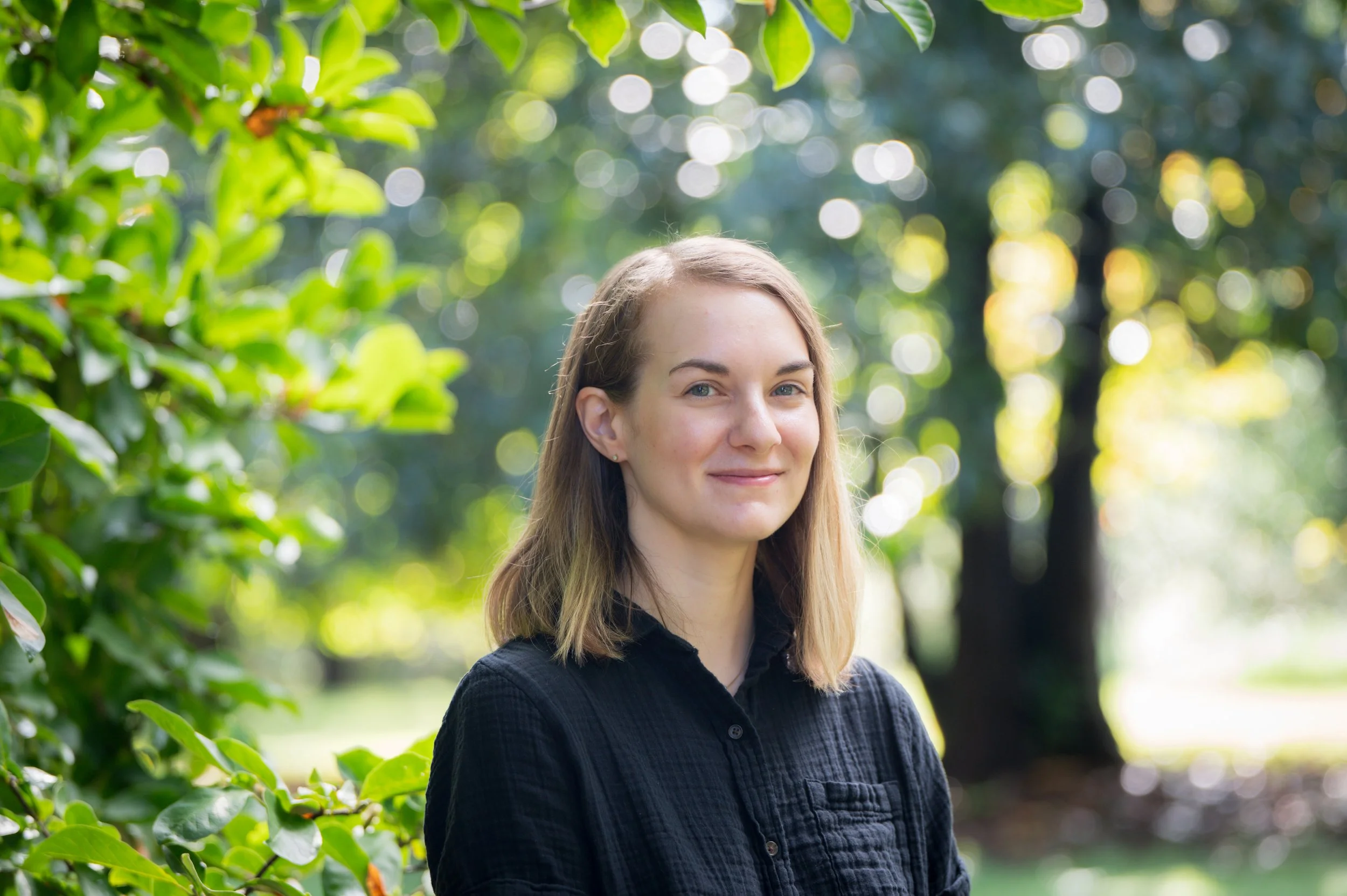 A young woman with shoulder-length light brown hair, wearing a black shirt, smiling softly outdoors among green foliage with sunlight filtering through trees in the background.