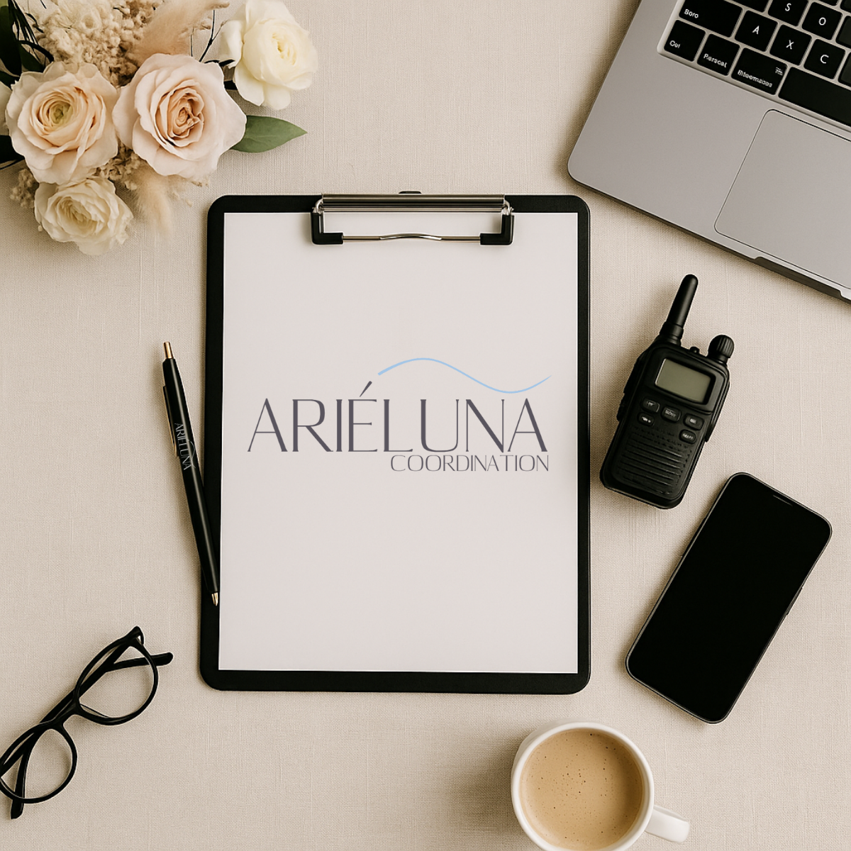 Desk setup with a bouquet of white and cream roses, a pen, glasses, a clipboard with Arieluna Coordination's logo, a walkie-talkie, a smartphone, a laptop, a cup of coffee, all arranged on a beige surface.