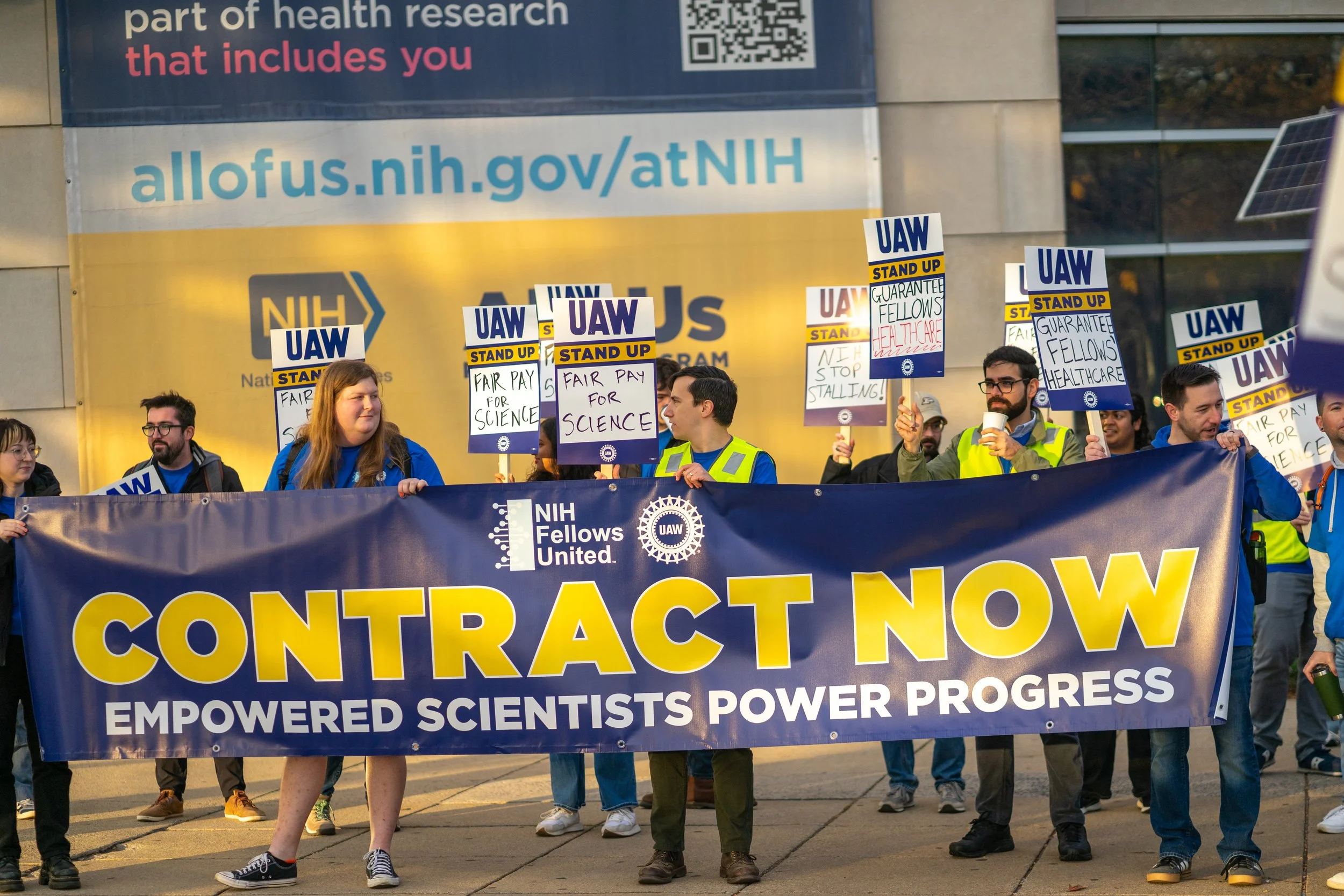 NIH Fellows holding a large Contract Now banner and UAW signs outside an NIH building.