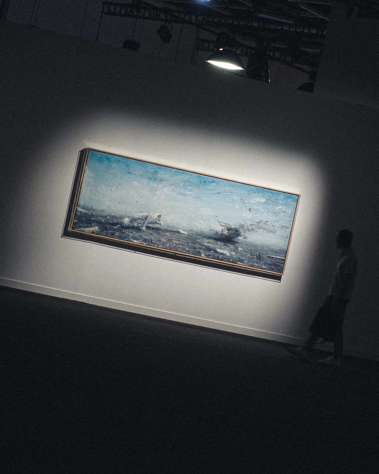 A person walking in an art gallery with a large landscape painting of a seascape illuminated on the wall.