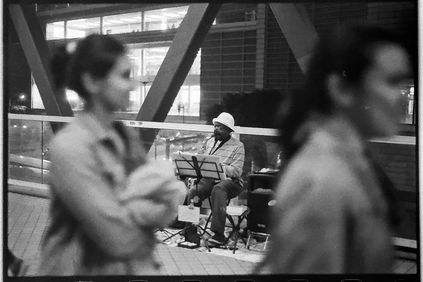 A black and white photo of a jazz musician playing a saxophone, seated outside a building with large glass windows. Two women are walking past in the foreground, slightly blurred.