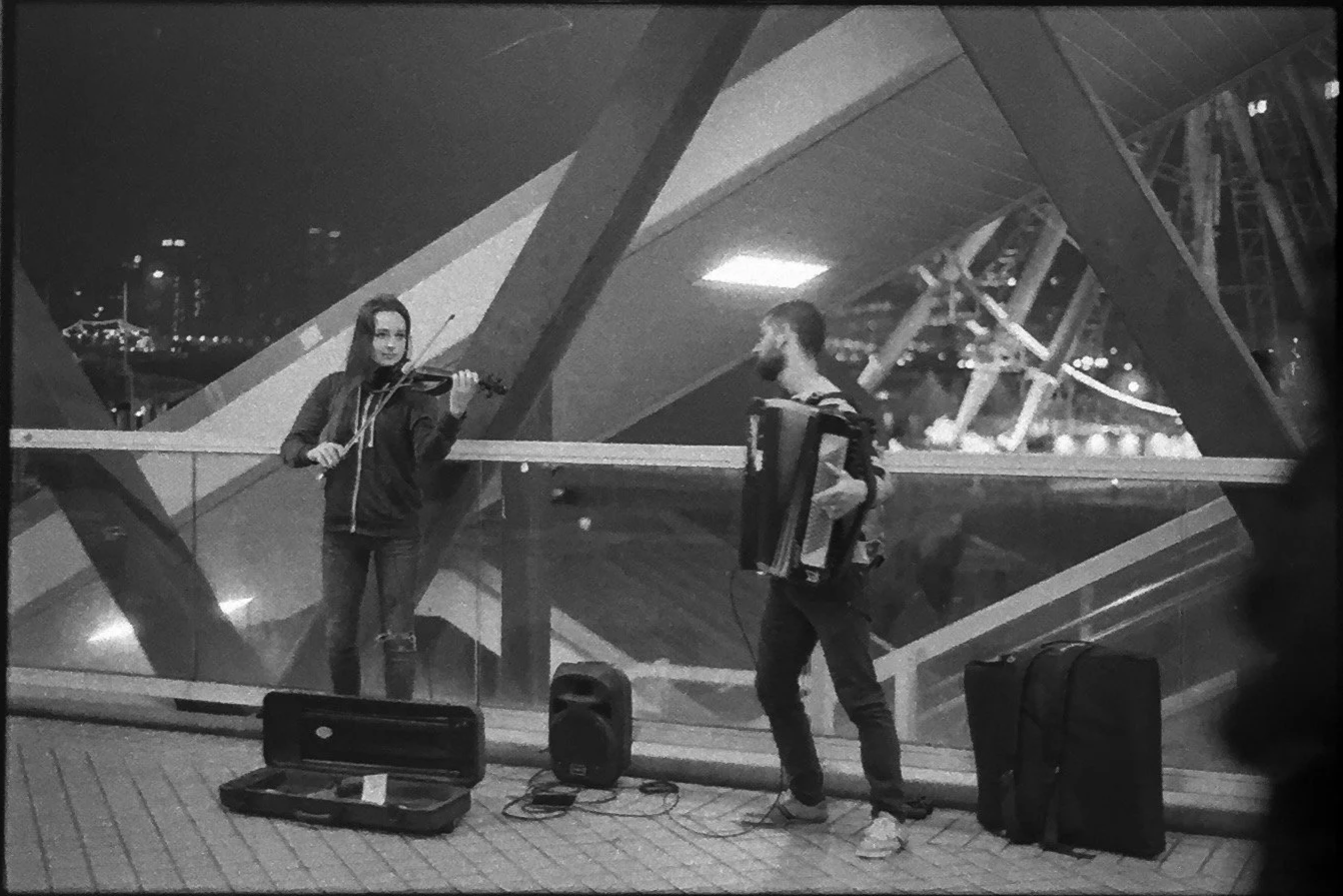 Two street musicians playing instruments inside a glass building at night, one with a violin and the other with an accordion, with open instrument cases and speakers on the ground in front of them.