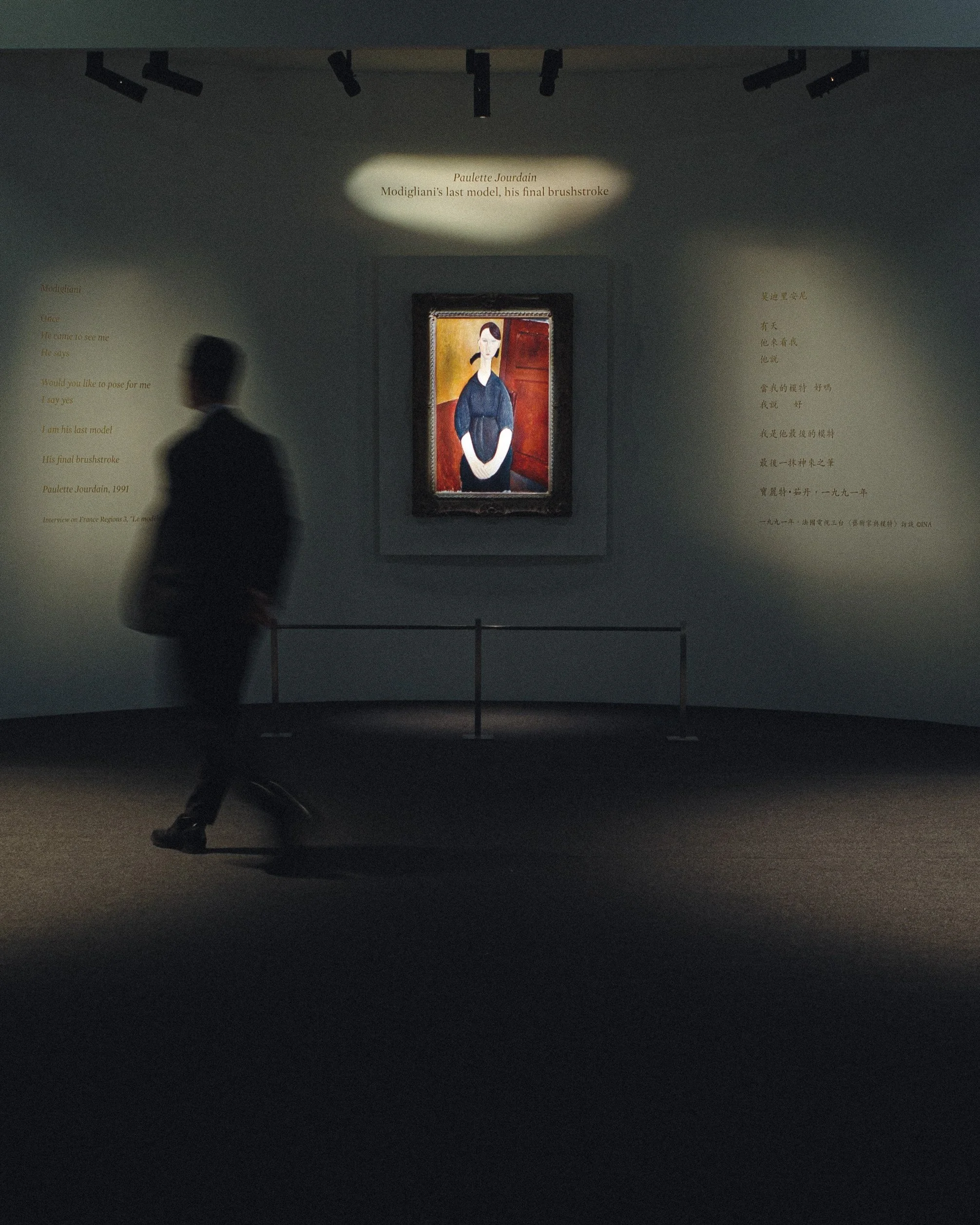 A person in a dark suit standing in a dimly lit art gallery, looking at a framed painting of a woman with dark hair tied back, wearing a dark blue dress, standing against a red and yellow background.