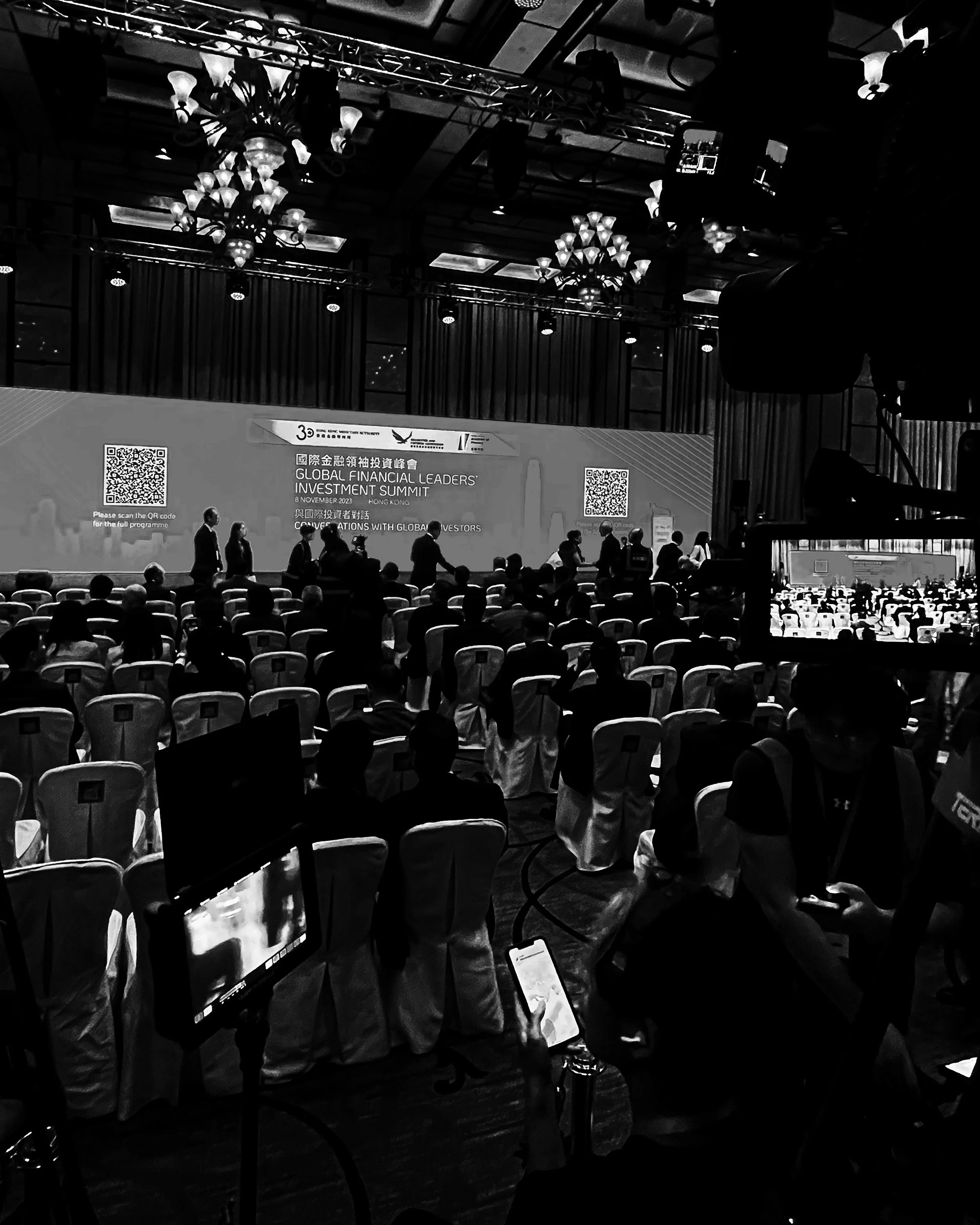 A large conference hall with many people attending the Global Financial Leaders Investment Summit in Hong Kong. The stage displays event information and QR codes. Attendees are seated, and some are using their phones, with professional equipment visible.
