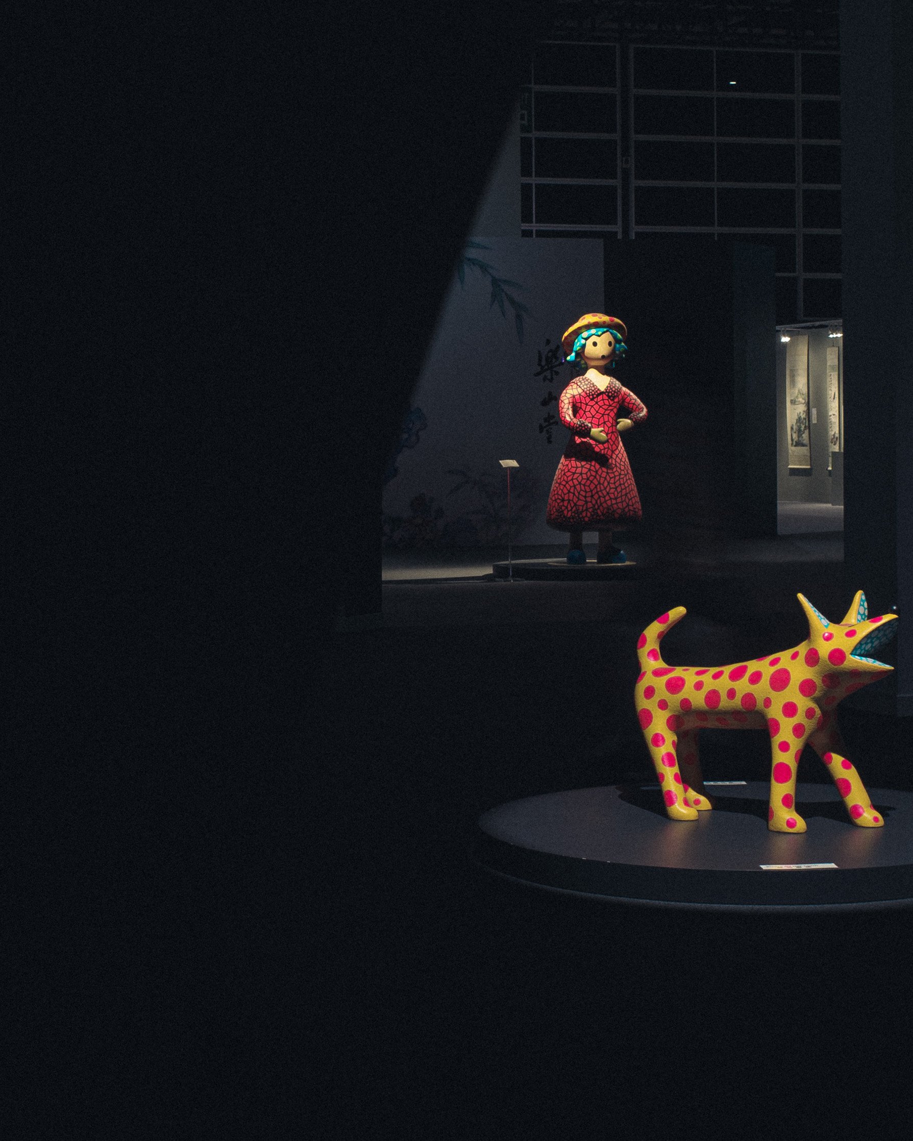 Colorful art sculptures of a woman in a red dress and a dotted pink and yellow dog in a dark exhibition space.