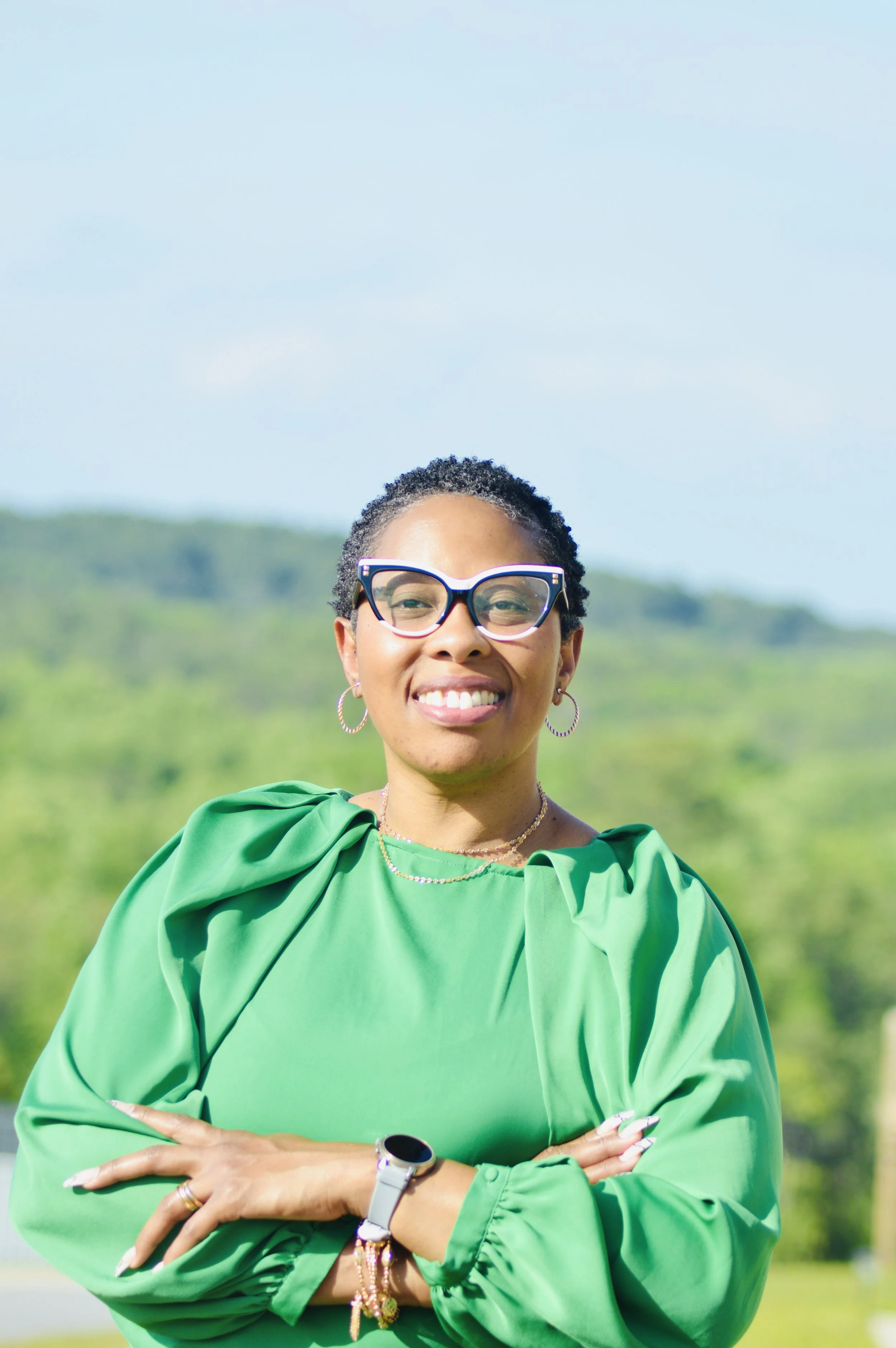 A woman with short curly hair, glasses, and hoop earrings smiling outdoors. She is wearing a bright green blouse with puffed shoulders and has her arms crossed. The background shows a lush, green landscape under a clear blue sky.