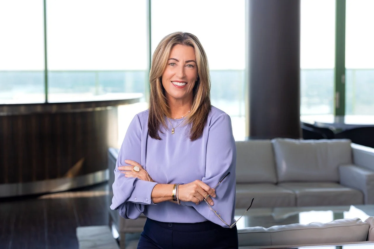 Marlo Zarka, smiling with shoulder-length light brown hair, wearing a lavender blouse, holding glasses, standing in a modern office lounge with large windows and a city view.