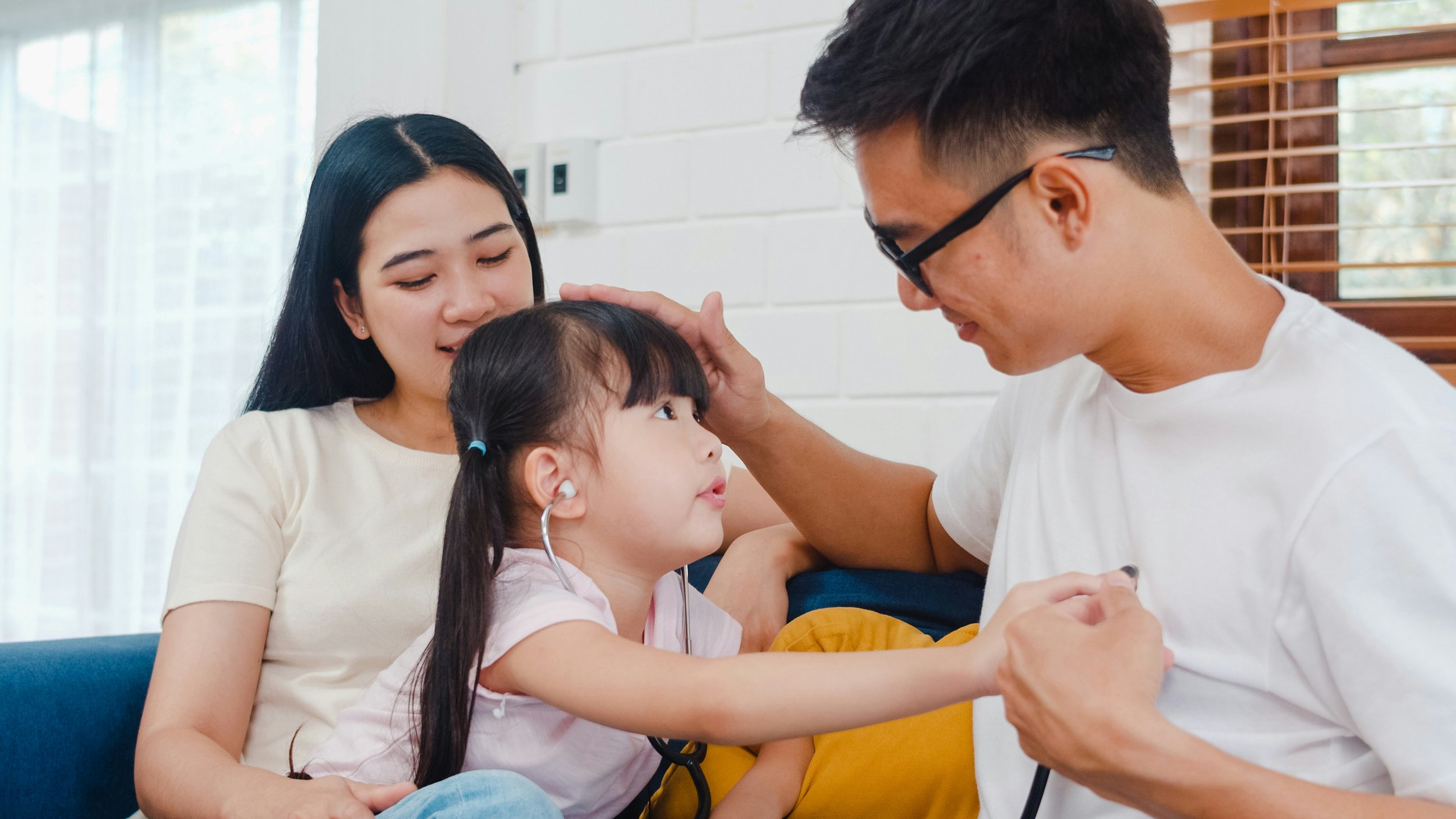 A young girl with a stethoscope listening to a man's chest while his mother gently touches his head in a bright living room.