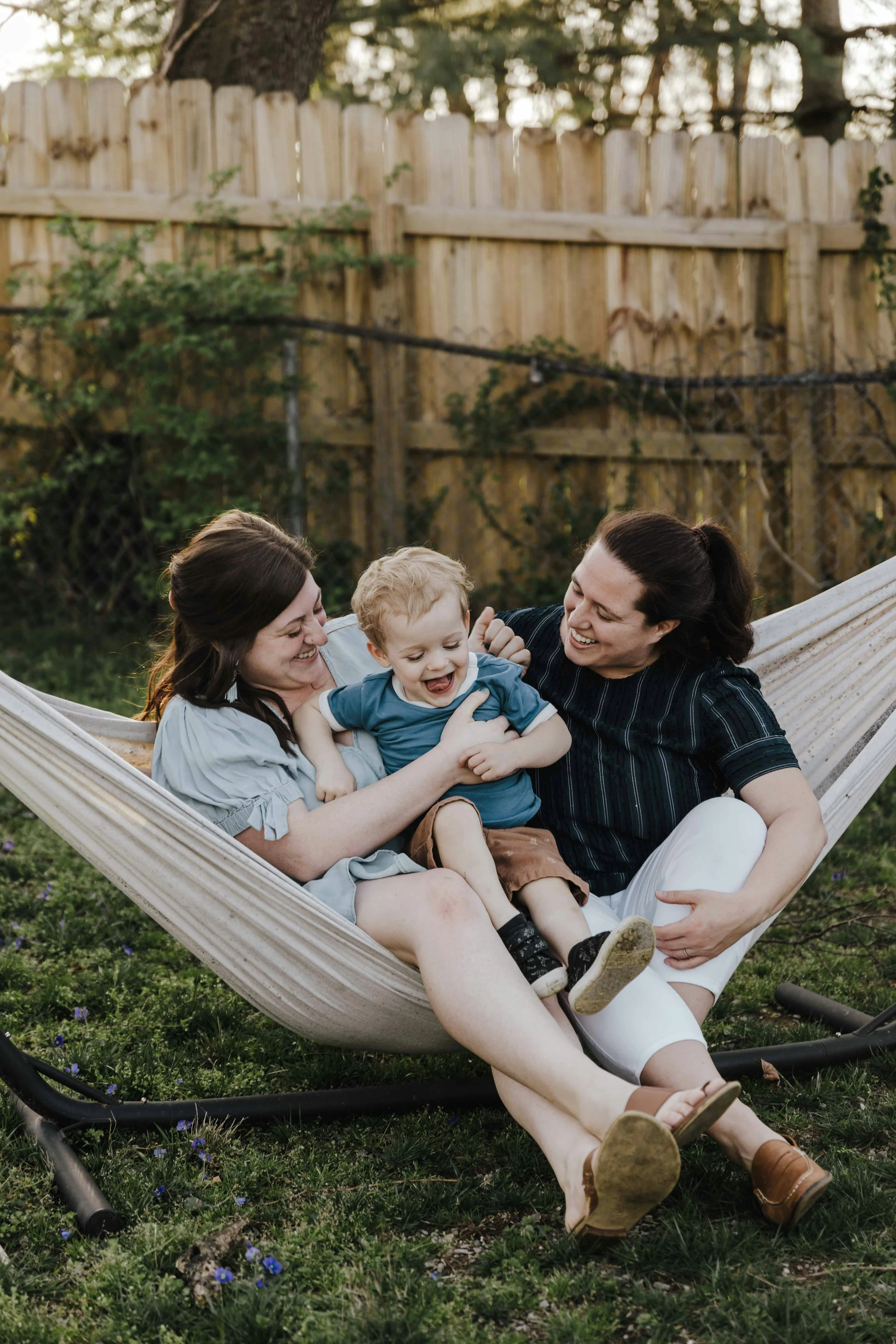 Two women and a young boy playing and laughing together on a hammock in a backyard.