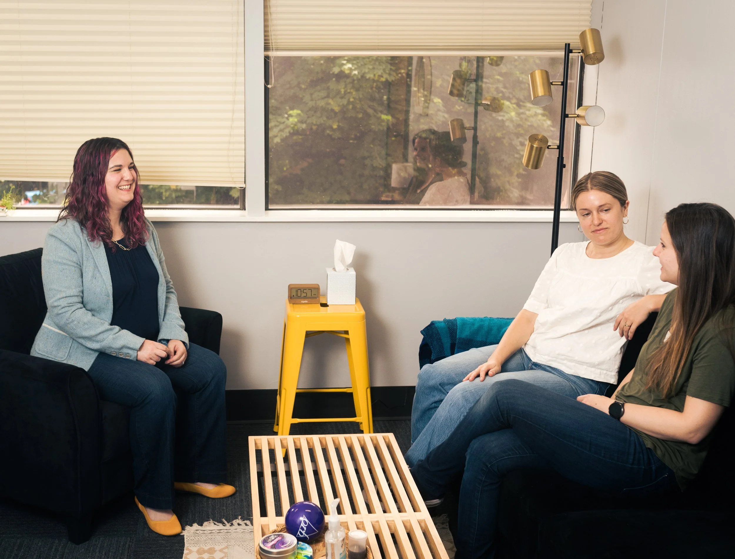 Three women having a conversation in a waiting room or casual office. One woman with purple and pink hair is smiling, sitting on a black sofa. The other two women are seated on another sofa, listening. There is a yellow table with tissues and a clock in the background, along with a large window showing greenery outside.