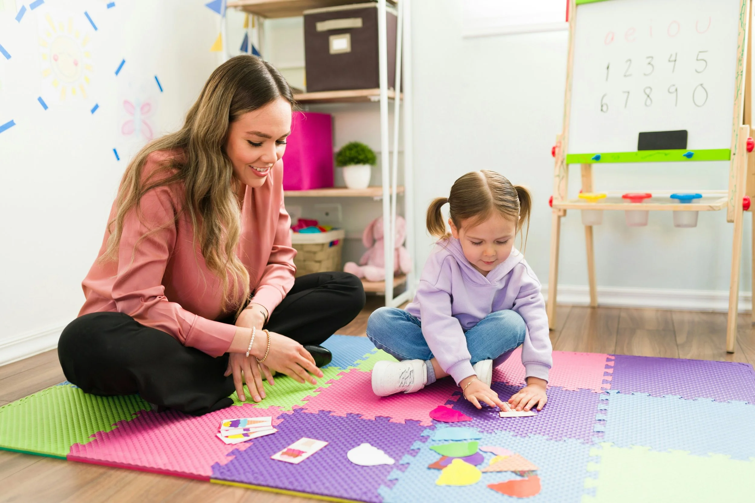 A woman and a young girl playing with colorful paper shapes on a foam mat in a classroom or playroom.