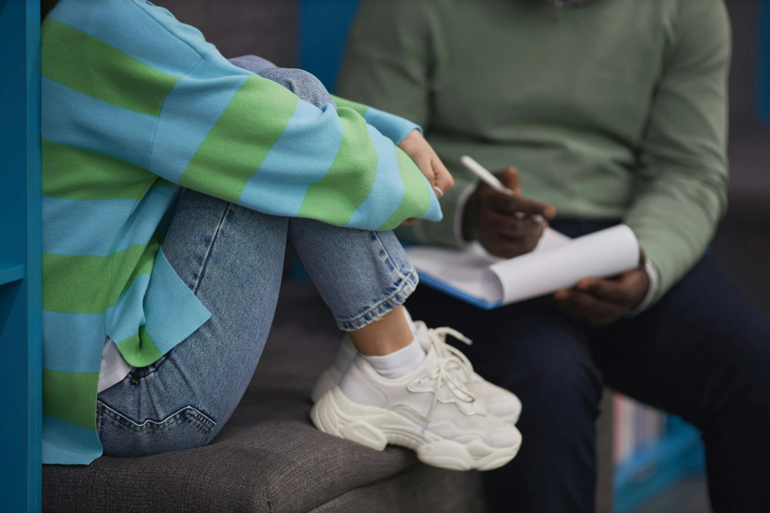 A young person sitting on a gray couch with their knees pulled up, wearing a colorful striped sweater, jeans, and white sneakers. A second person beside them is holding a clipboard and writing with a pen.