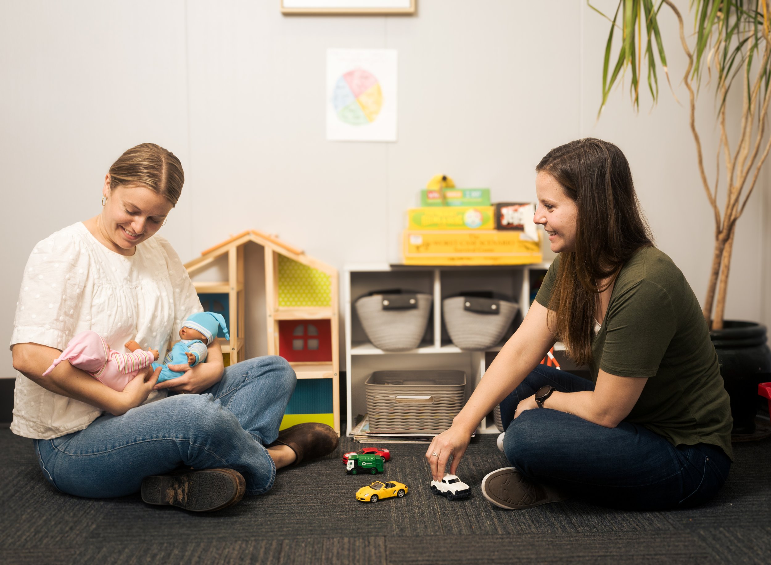 Two women sitting on the floor in a playroom, with the woman on the left holding a baby, and the woman on the right playing with toy cars.