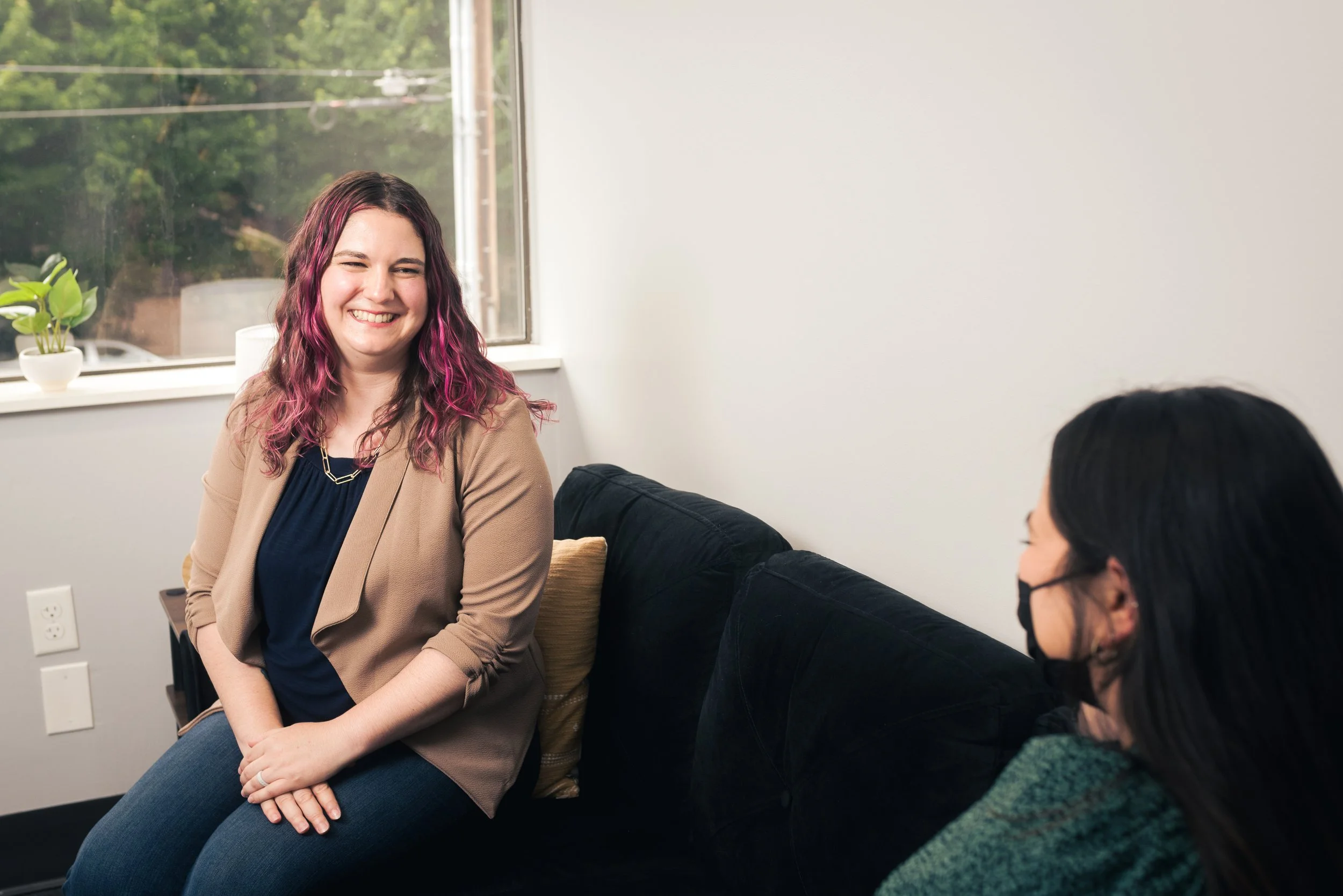 Two women sitting and talking in a bright room with a window and a small potted plant.