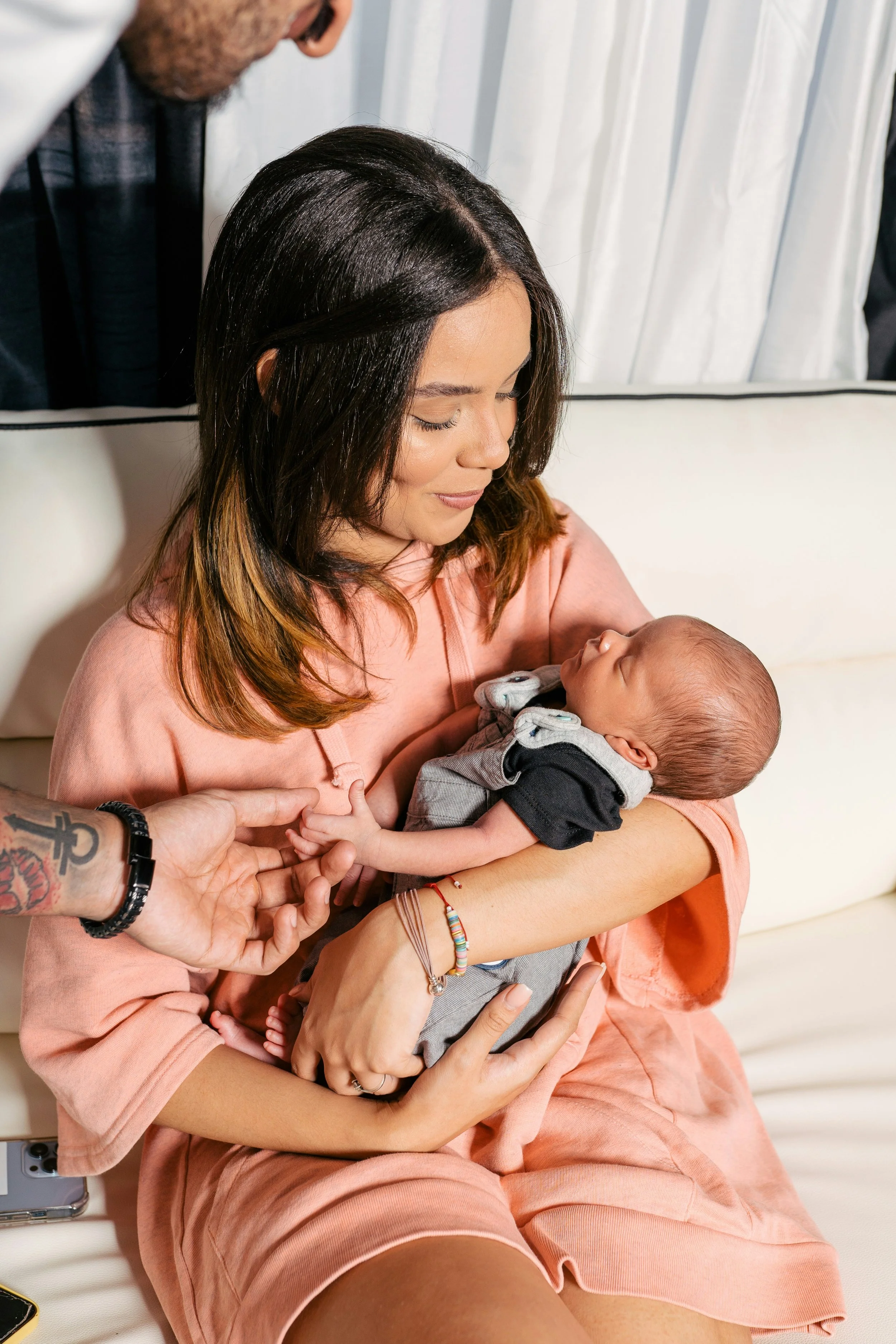 A woman with brown hair and a pink dress holding a newborn baby while someone adjusts the baby's hand.