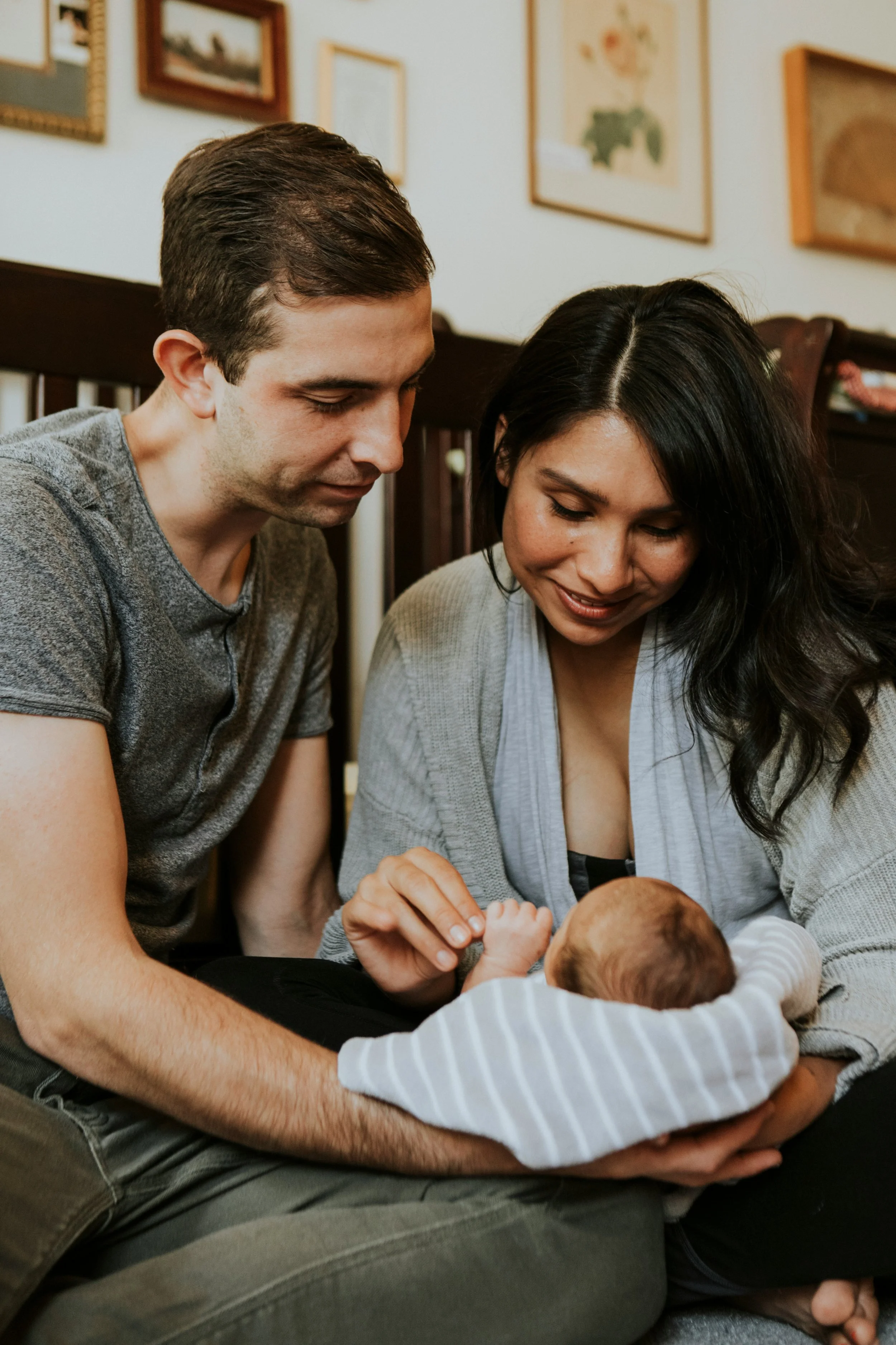 A family of three, a man, woman, and a baby, sitting together in a cozy room, holding and looking at their baby with love.