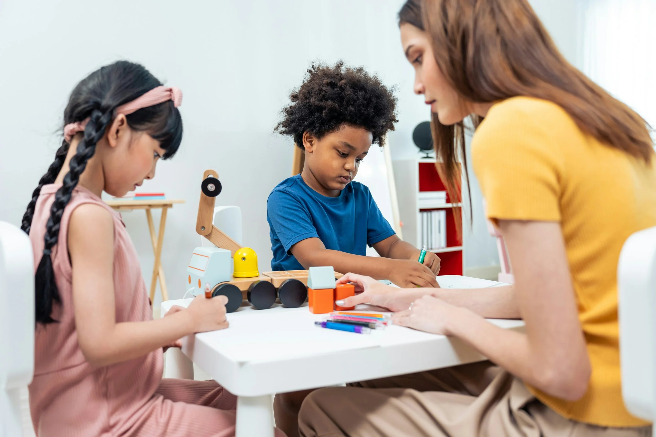 A woman with red hair in a yellow shirt teaches two young children, a girl and a boy, how to build a robot with wooden and plastic parts and colorful markers at a white table.