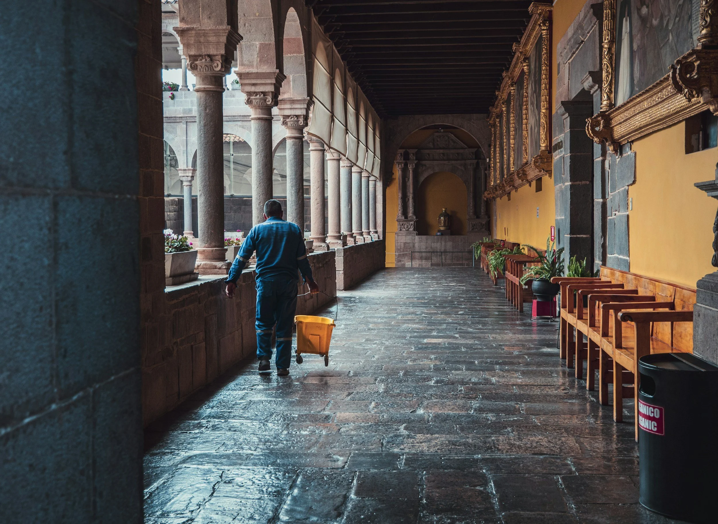 A man walking through an arched corridor with stone columns and a wet, reflective floor, pushing a yellow cleaning cart.