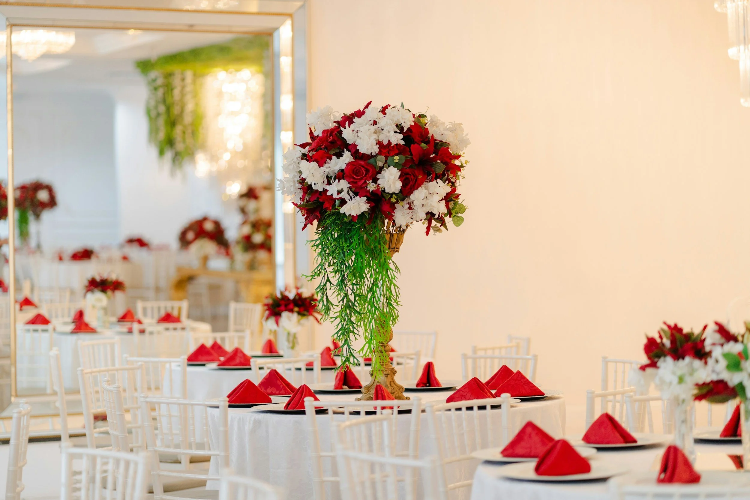 Wedding or event reception decorated with red and white floral centerpieces, set tables with red napkins, and white chairs in a bright, elegant room.