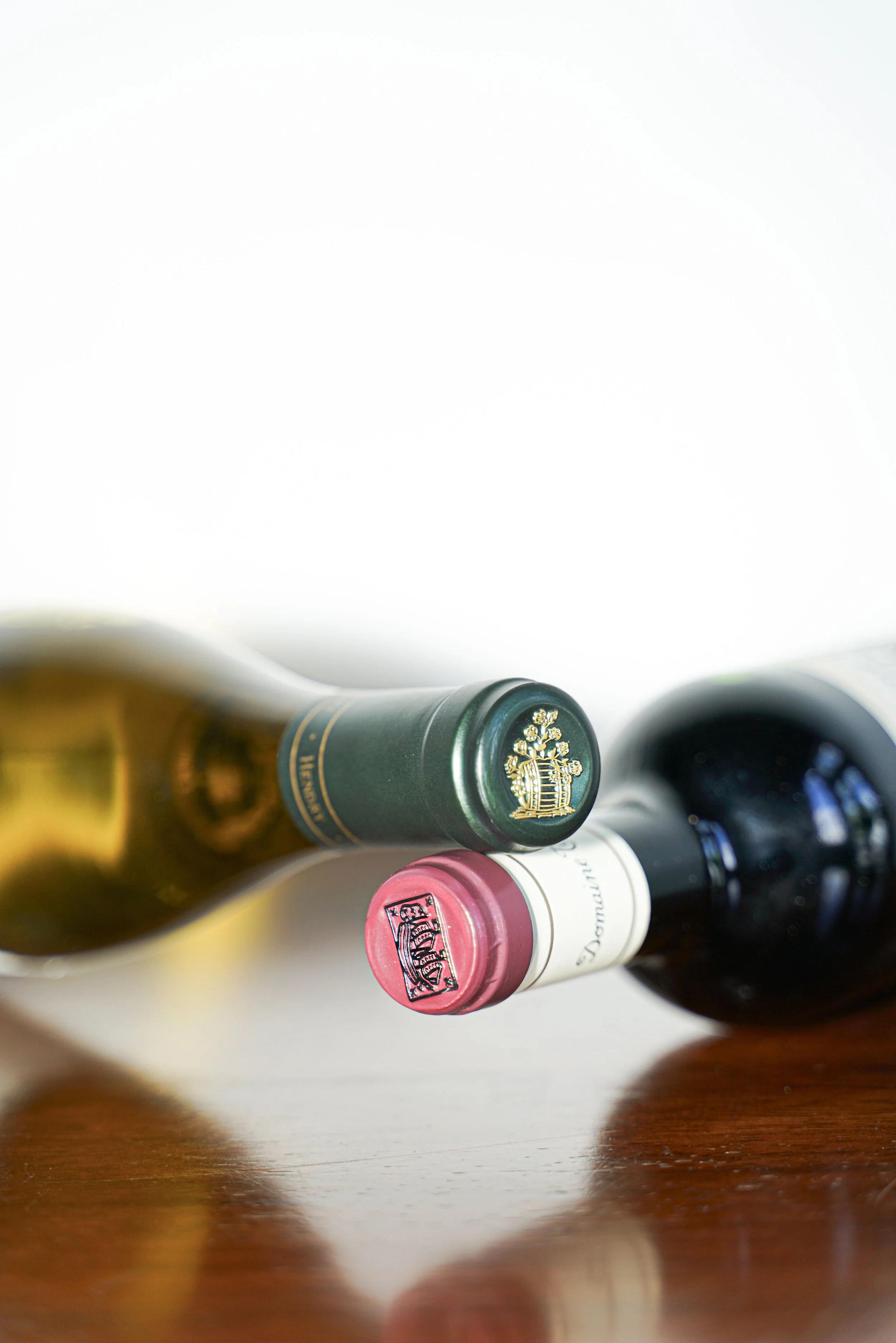 The image shows the necks of two wine bottles lying on a wooden surface, with one golden colored bottle on the left and a dark color bottle on the right. The labels are partially visible.