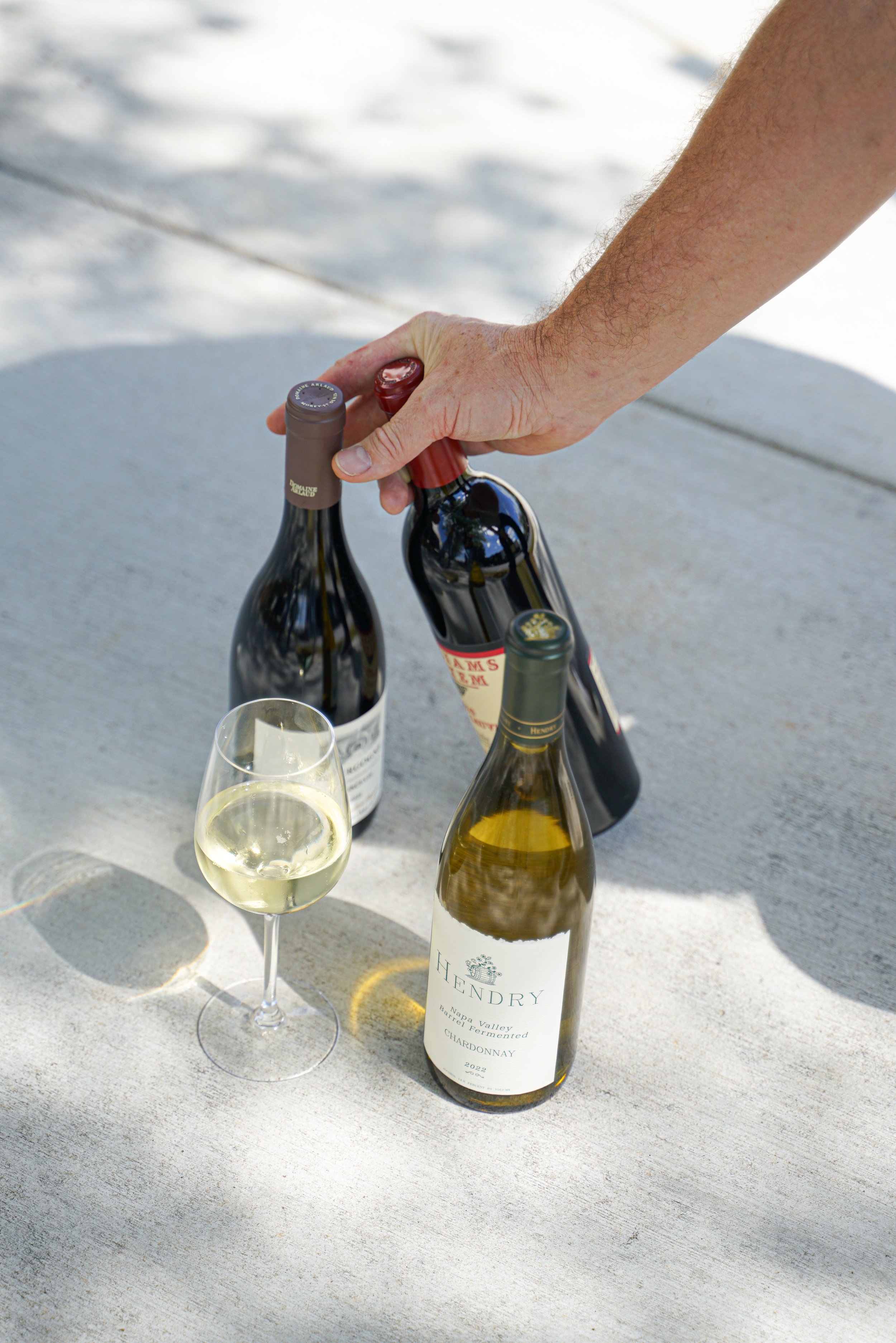 Person's hand opening a bottle of white wine on an outdoor table with two other wine bottles and a filled wine glass.