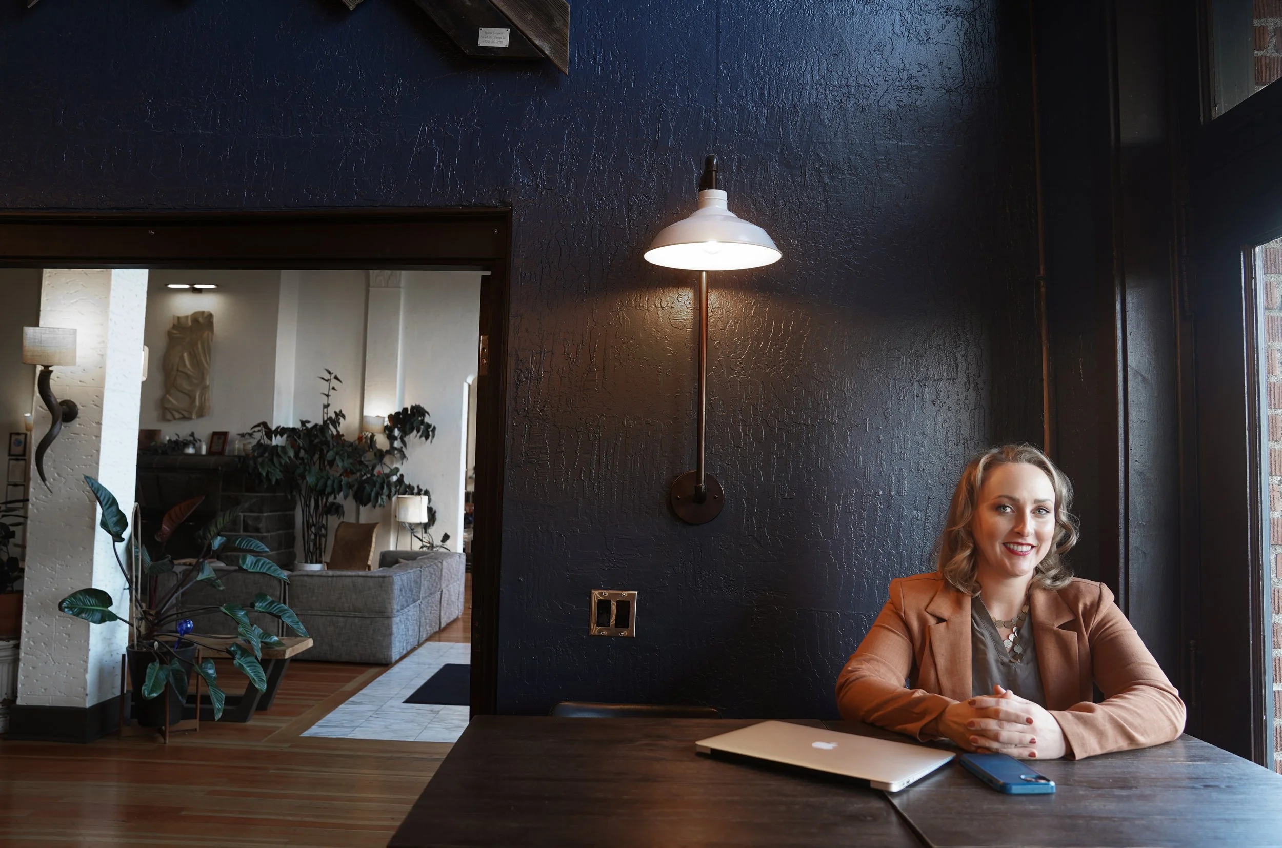 Diana Pullen Real Estate Broker in Redmond, Oregon in Central Oregon in brown blazer sitting at a table with a laptop and smartphone in a cafe with dark walls and window.