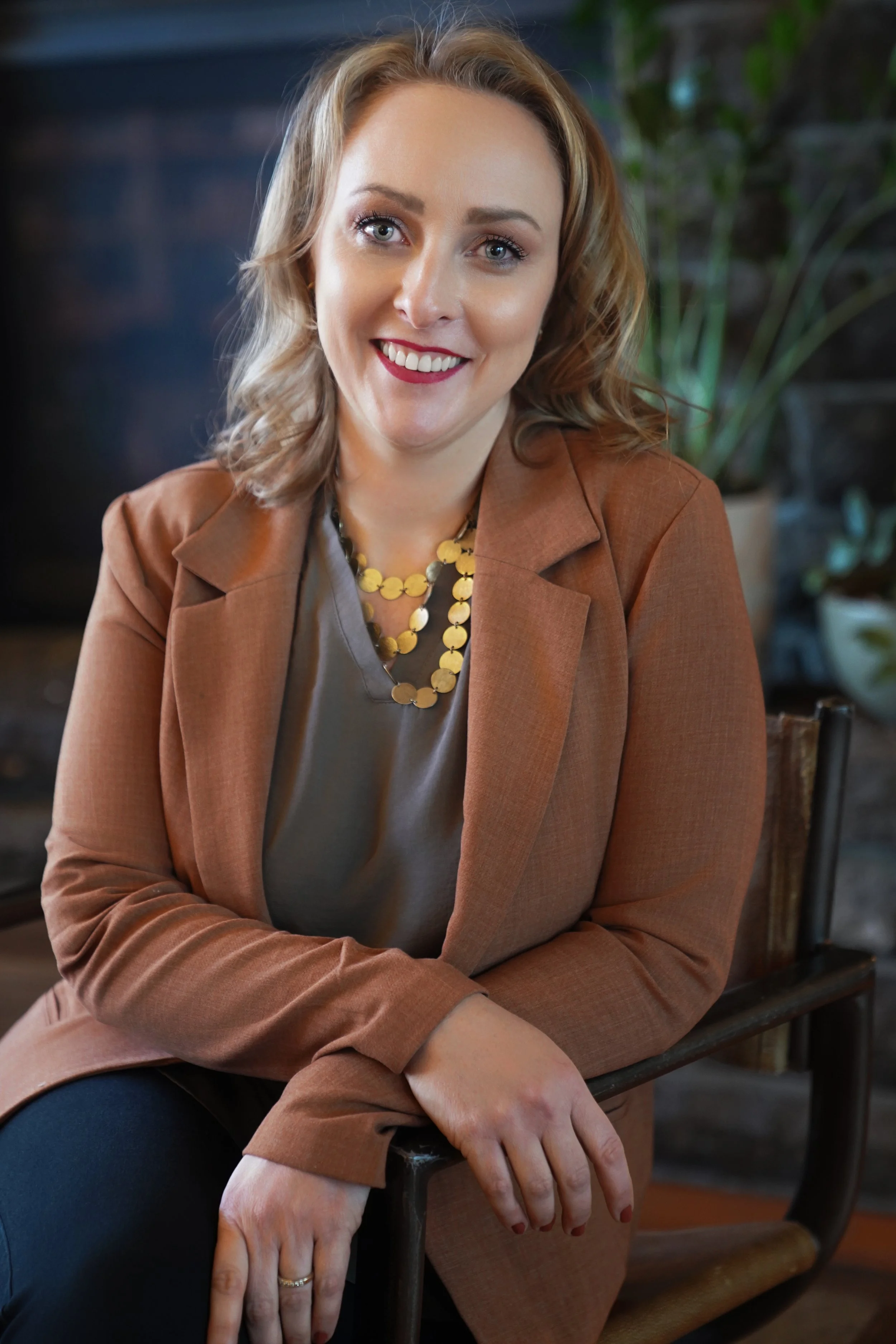 Diana Pullen Real Estate Broker in Redmond, Oregon in Central Oregon, wearing a brown blazer, gold necklace, and makeup, sitting indoors with a plant and dark wall in the background.