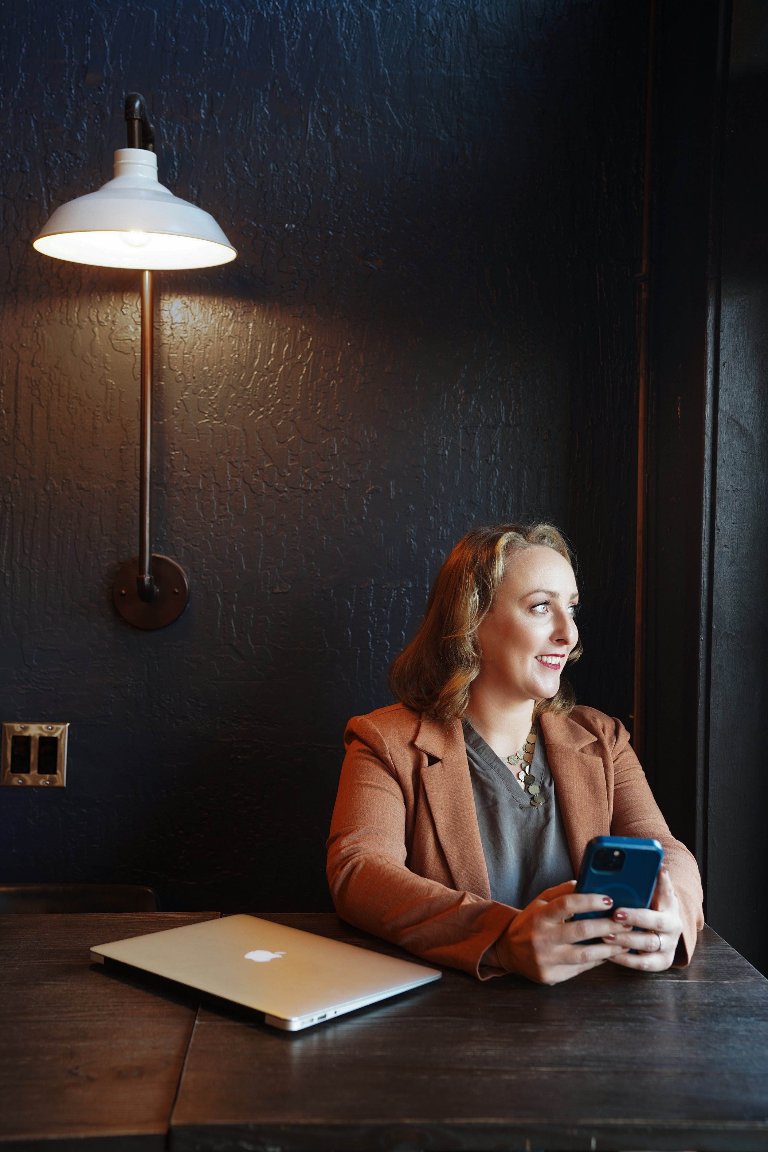 Diana Pullen Redmond Oregon real estate broker sitting at a dark wooden table in a dimly lit room, holding a blue phone and smiling, with a laptop and closed notebook on the table.