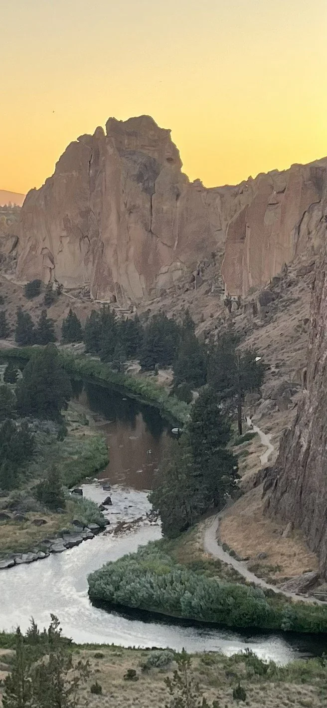 Smith Rock at sunset featuring a river running through a canyon with tall rocky cliffs and dense green trees along the riverbank.