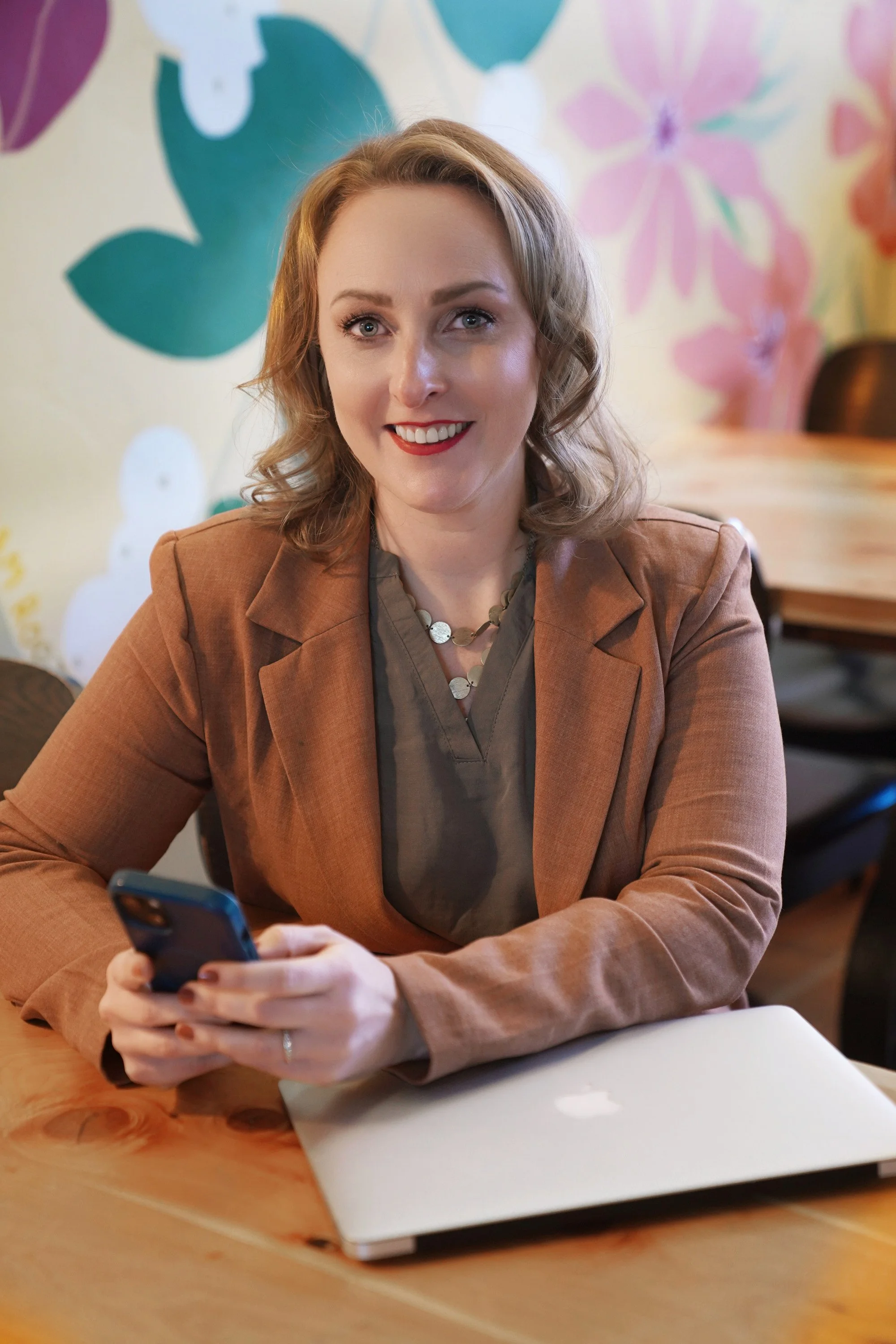 Diana Pullen, Redmond Oregon real estate broker  in Central Oregon, sitting at a wooden table with a silver laptop, a smartphone, and papers. She is smiling and looking at the camera, with colorful floral wall art in the background.