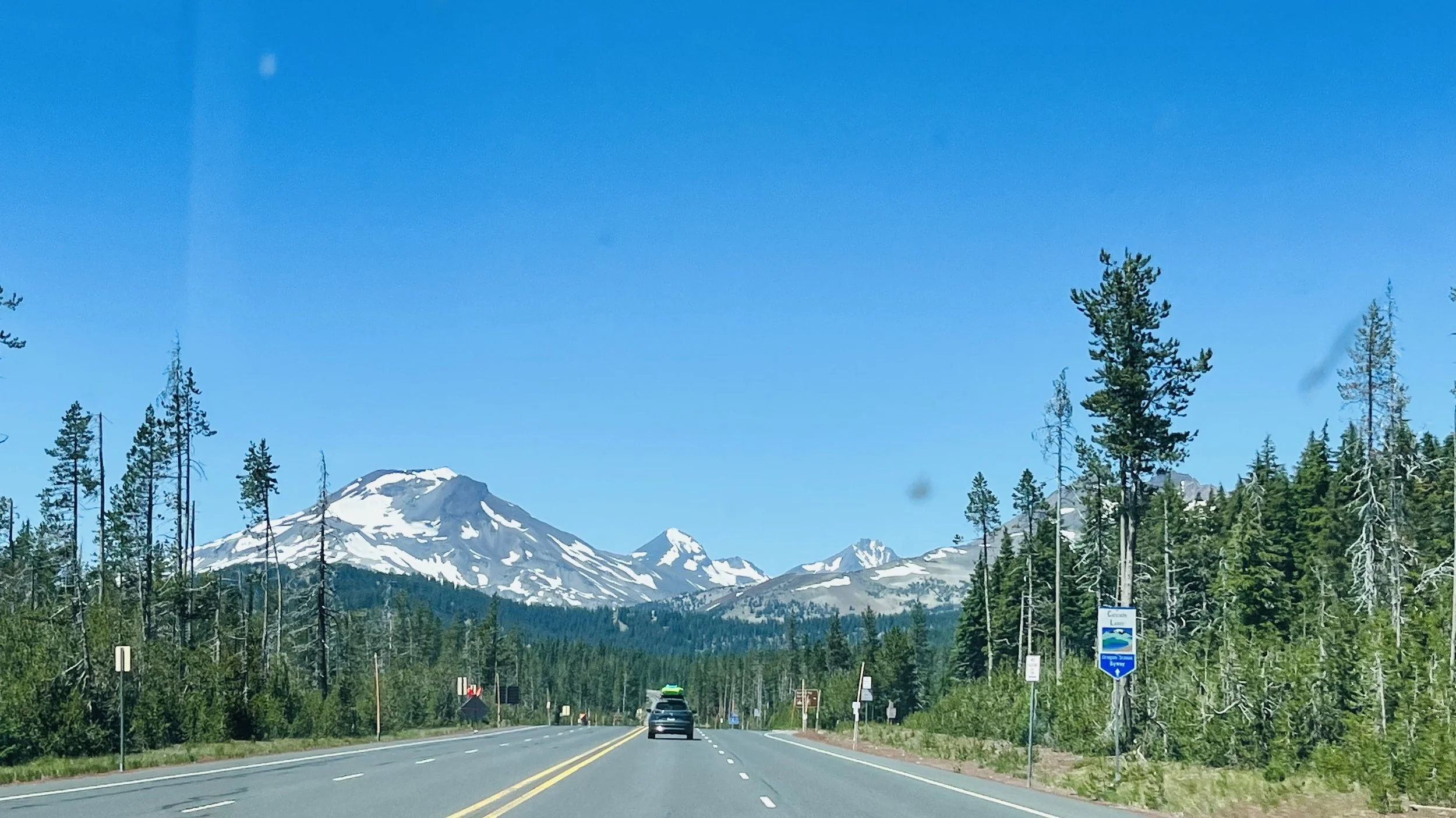 Highway 20 leaving Sisters, Oregon with cars driving past tall trees and snow-capped mountains under a bright blue sky.
